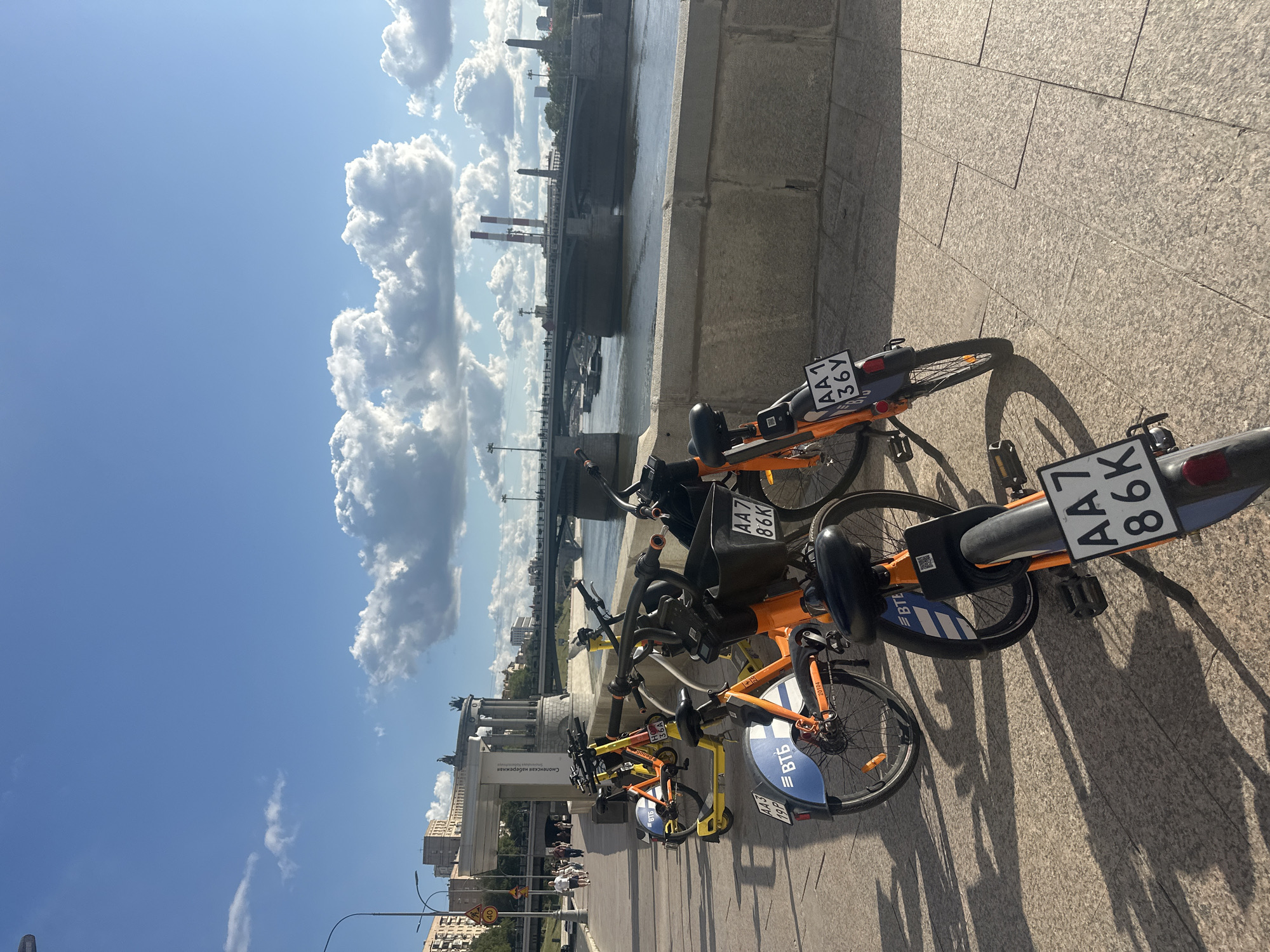 The image shows a row of rental bicycles parked on a paved embankment next to a wide river, likely the Moskva River in Moscow, Russia. The sky is blue with fluffy white clouds, and the sun is casting long shadows from the bicycles onto the pavement, indicating it is either late morning or late afternoon.
In the foreground, several orange and yellow rental bicycles are visible. One bicycle in the very front has a license plate that reads "AA7 86K". Behind it, another bicycle has a license plate "AA1 36Y" and the "ВТБ" logo. Further back, a yellow bicycle and another orange bicycle are parked in a row.
The embankment has a stone wall bordering the river. Across the river, a large bridge spans the water, with traffic visible on it. In the background, there are buildings, including apartment blocks and a structure with prominent columns and statues, possibly a monument or government building. Tall industrial chimneys with red and white bands are also visible in the distance, along with other cityscape elements. A few people are walking on the embankment in the middle ground, further away from the parked bicycles. The overall scene depicts a typical urban landscape with recreational and transportation elements integrated into the city's infrastructure.