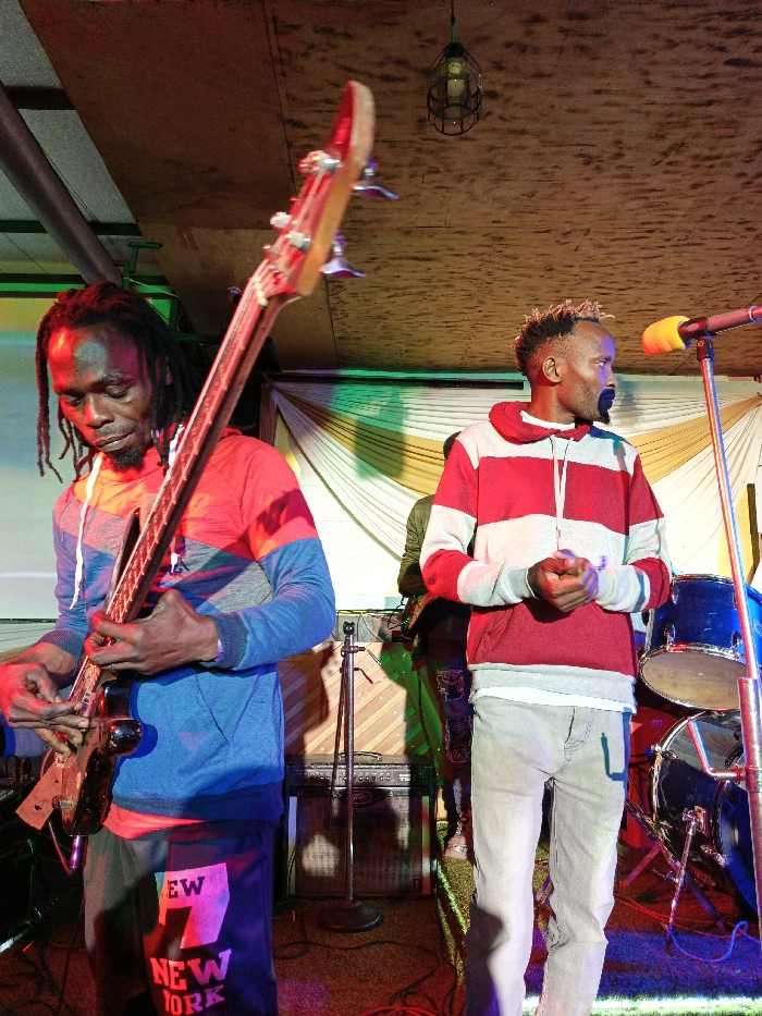 The image depicts an indoor live music performance featuring two male musicians at a venue in Nairobi, Kenya. On the left, a dark-skinned male with dreadlocks, wearing a red, grey, and blue hooded sweatshirt and black shorts with "NEW YORK" lettering, plays a black bass guitar. His gaze is directed downwards at the instrument as both hands manipulate the strings and fretboard. To the right stands a male with lighter skin, short bleached hair, and a prominent dark goatee, dressed in a red and grey horizontally striped hooded sweatshirt over a white undershirt and light-colored jeans. His hands are clasped together at waist level, and he looks towards the viewer's left.
The scene is illuminated by red, blue, and green stage lighting. Visible stage equipment includes a microphone with an orange foam cover on a stand, components of a blue drum kit, and an amplifier system positioned behind the bassist. The background features draped, pleated fabric, predominantly white with yellow accents, serving as a stage backdrop. The ceiling is constructed from wooden panels, with a single caged light fixture suspended overhead. The floor appears dark, reflecting the colored stage lights.