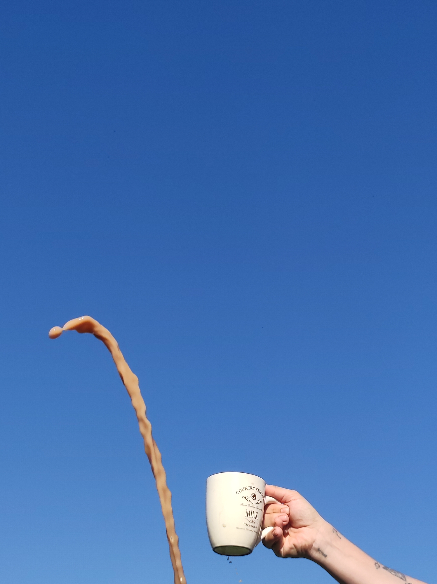 A person holds a white mug with "Country Kitchen, Fresh Daily, MILK" printed on it in front of a clear blue sky. A stream of tan-colored liquid is pouring out of the mug. The person's arm is visible, and there are tattoos on their wrist and forearm. The location is not indicated in the image.