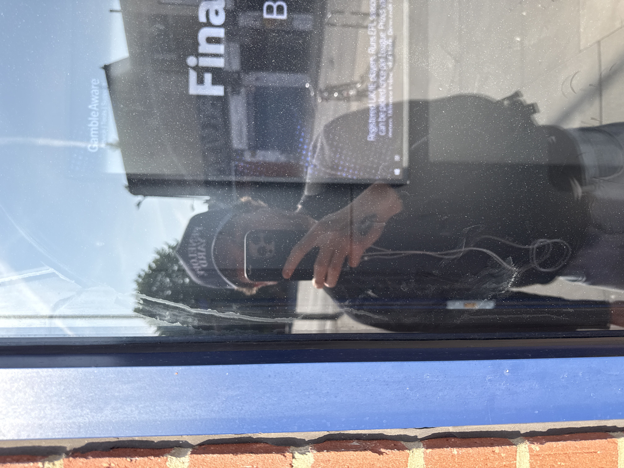 The image shows a reflection in a dusty shop window. The reflection includes a person taking a photo with a smartphone. The person is wearing dark clothing and a hat. The window displays text that reads "Gamble Aware", "Fina", "B", and information related to UK/IE players and EPL prizes. Below the window is a blue painted surface and a brick wall. The location appears to be a commercial area. There is no specific information about location in the city of London, United Kingdom.