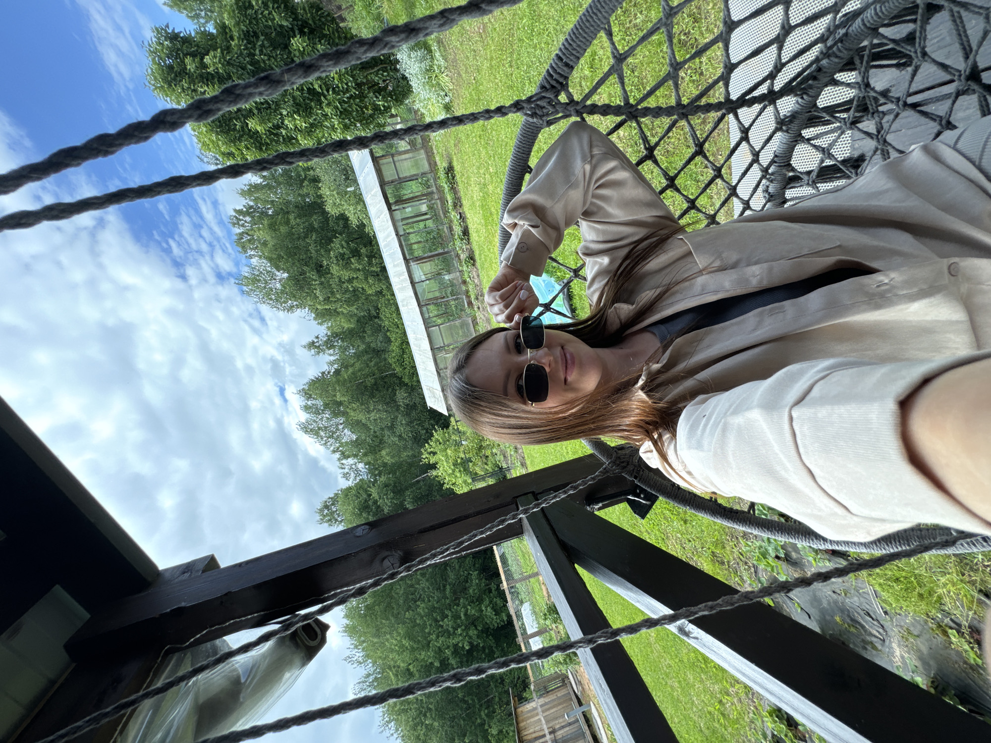 A young woman with long brown hair and sunglasses is sitting in a hanging chair on a wooden porch in a rural setting. She is wearing a light-colored shirt. The background shows a green lawn, trees, a greenhouse, and a wooden structure. The sky is cloudy with some blue patches. The image appears to be taken in the city of Ponazyrevo, Russia, given the context of the provided crops.