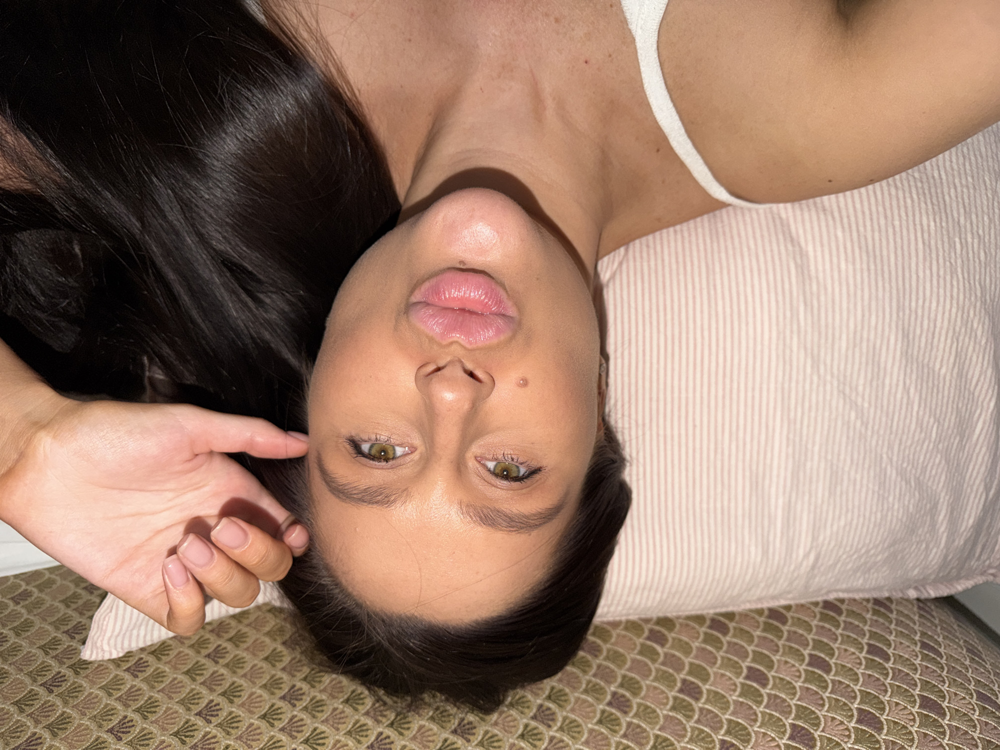 A medium shot depicts a woman lying down, viewed from an inverted perspective. Her head rests on a pink-striped pillow, which in turn is positioned on a patterned surface. The woman's dark hair is prominently displayed. Her right hand is raised near her face. She has a white tank top on. There is no indication of this image being taken in Khimki, Russia.