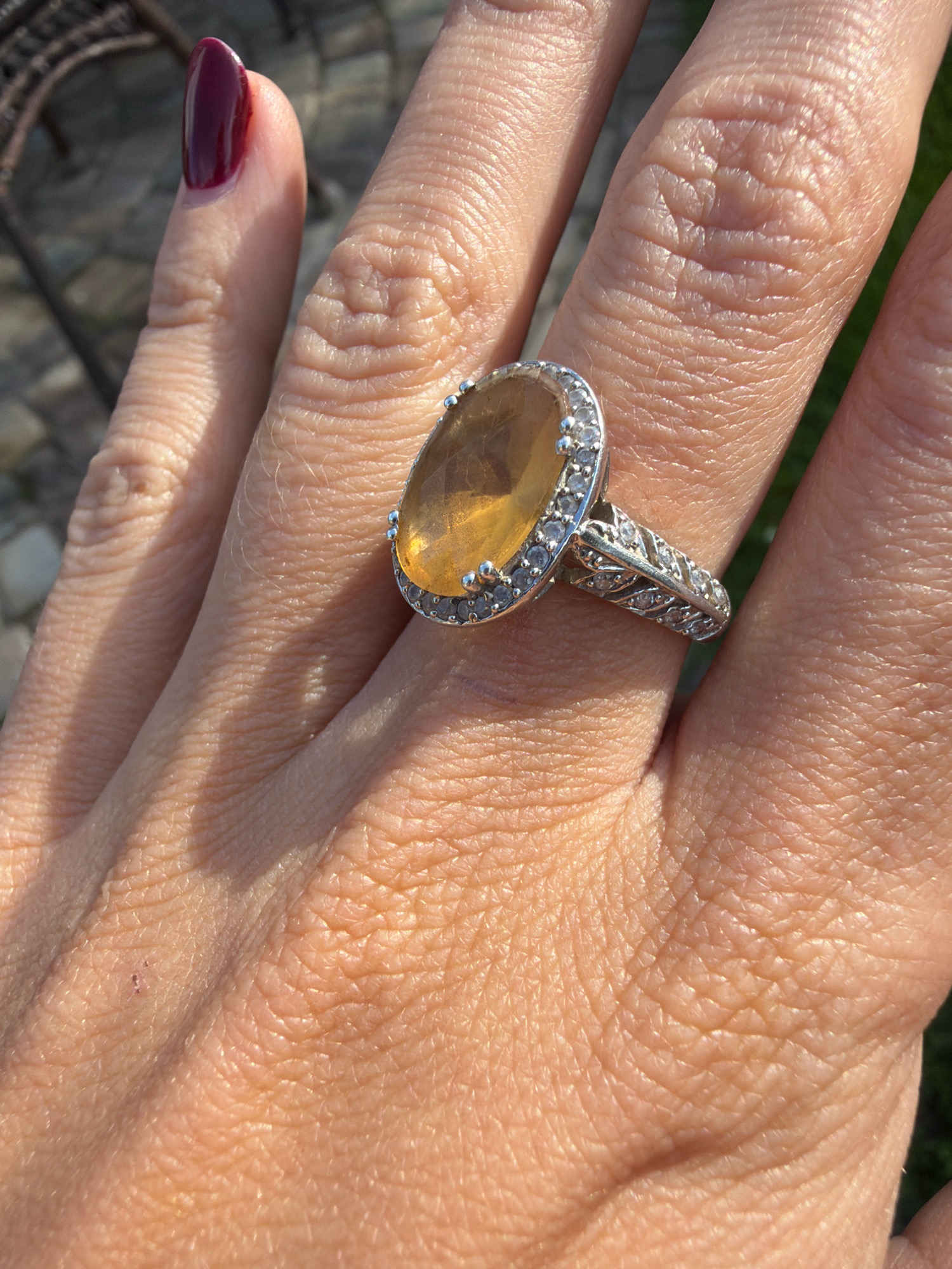 A close-up, over-the-shoulder shot shows a woman's hand adorned with a silver ring featuring a large oval yellow gemstone surrounded by small diamonds. The ring is positioned on her ring finger, which has a fingernail painted in a dark burgundy color. The hand exhibits textured skin with visible wrinkles and fine hairs, illuminated by bright sunlight, casting a shadow. The background is blurred, suggesting an outdoor setting with hints of gravel and greenery, possibly indicating the outskirts of Бабініцкі сельскі Савет, Belarus.
