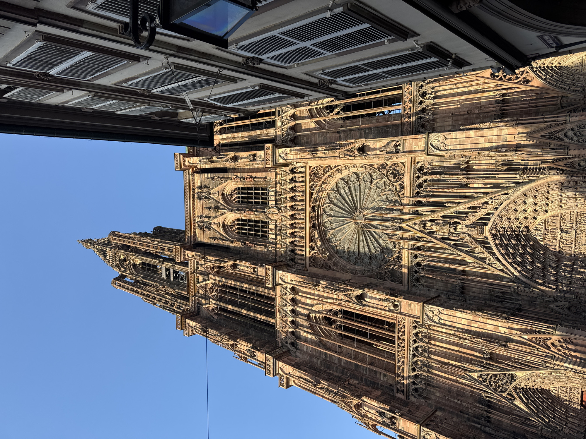 The image shows the Cathedral of Notre-Dame of Strasbourg in Strasbourg, France, taken from a low angle, looking upwards. The Gothic architecture of the cathedral is prominently displayed, including its rose window, spires, and intricate stone carvings. The building appears to be lit by sunlight from the side. The top of the image includes elements of a building across the street, with details of the architecture, including window shutters. The sky is clear and blue. A thin electrical wire is stretched across the image from the upper left corner.