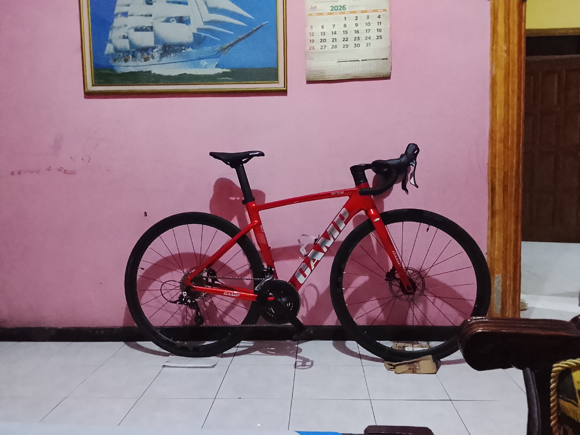 A red road bicycle with black wheels is parked against a pink wall. Above the bicycle, a framed painting of a sailing ship hangs on the wall, along with a calendar for July 2026. The bicycle has the word "GAMP" written on its frame. The floor is tiled with white and grey squares. To the right of the bicycle, a wooden pillar and a doorway are visible, suggesting the scene is indoors. In the foreground, a portion of a dark wooden chair with yellow and black trim is visible. The image appears to be taken in the city of Karangkemiri, Indonesia.