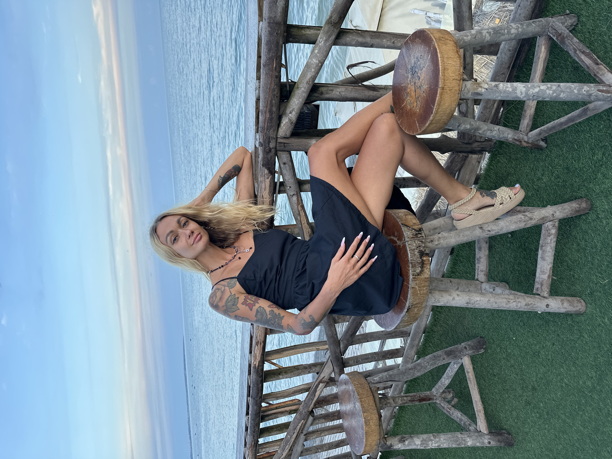 In Phu Thuy Ward, Vietnam, a young woman with blonde hair and numerous tattoos on her arms sits on a wooden stool at a table overlooking the sea. She wears a black sundress and beige sandals. The table is made of rough-hewn wood, as are the stools. The setting appears to be an outdoor restaurant or bar with a wooden railing and artificial turf. The sea stretches out behind her, with a pale blue sky and a few wisps of clouds. The image captures a moment of relaxation and enjoyment in a tropical setting.