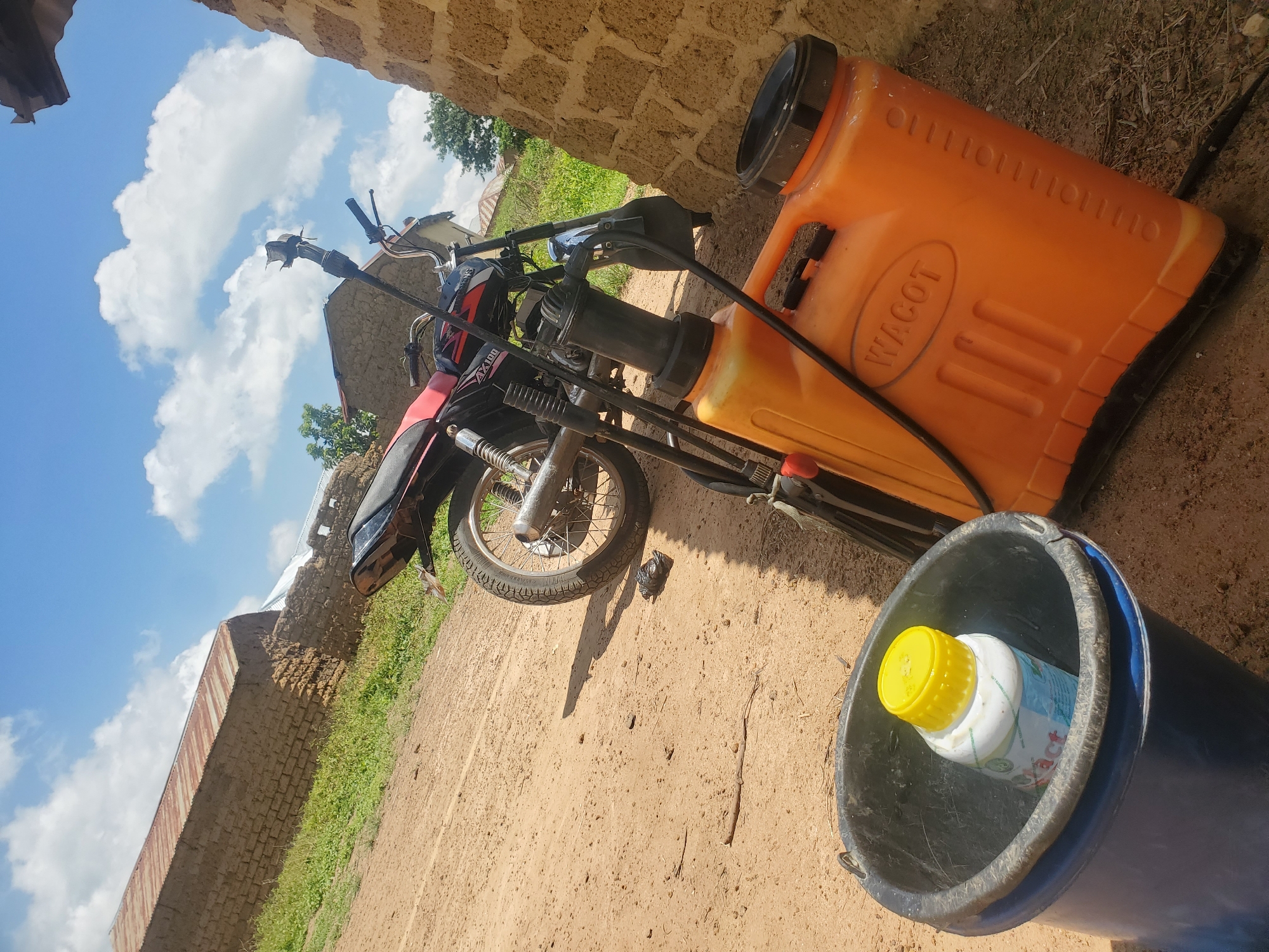 In Wumat Central, Nigeria, a scene depicts an outdoor rural environment with a dirt ground and a few buildings constructed from mud bricks. The sky is blue with scattered white clouds. In the foreground, a dark blue bucket contains a white bottle with a yellow cap. Next to the bucket is a bright orange liquid sprayer with a black hose and handle. Behind these items, a motorcycle is parked, facing to the left. The motorcycle has a red seat and black body. The rear wheel of the motorcycle is visible. A portion of a mud brick wall is on the right side of the image.