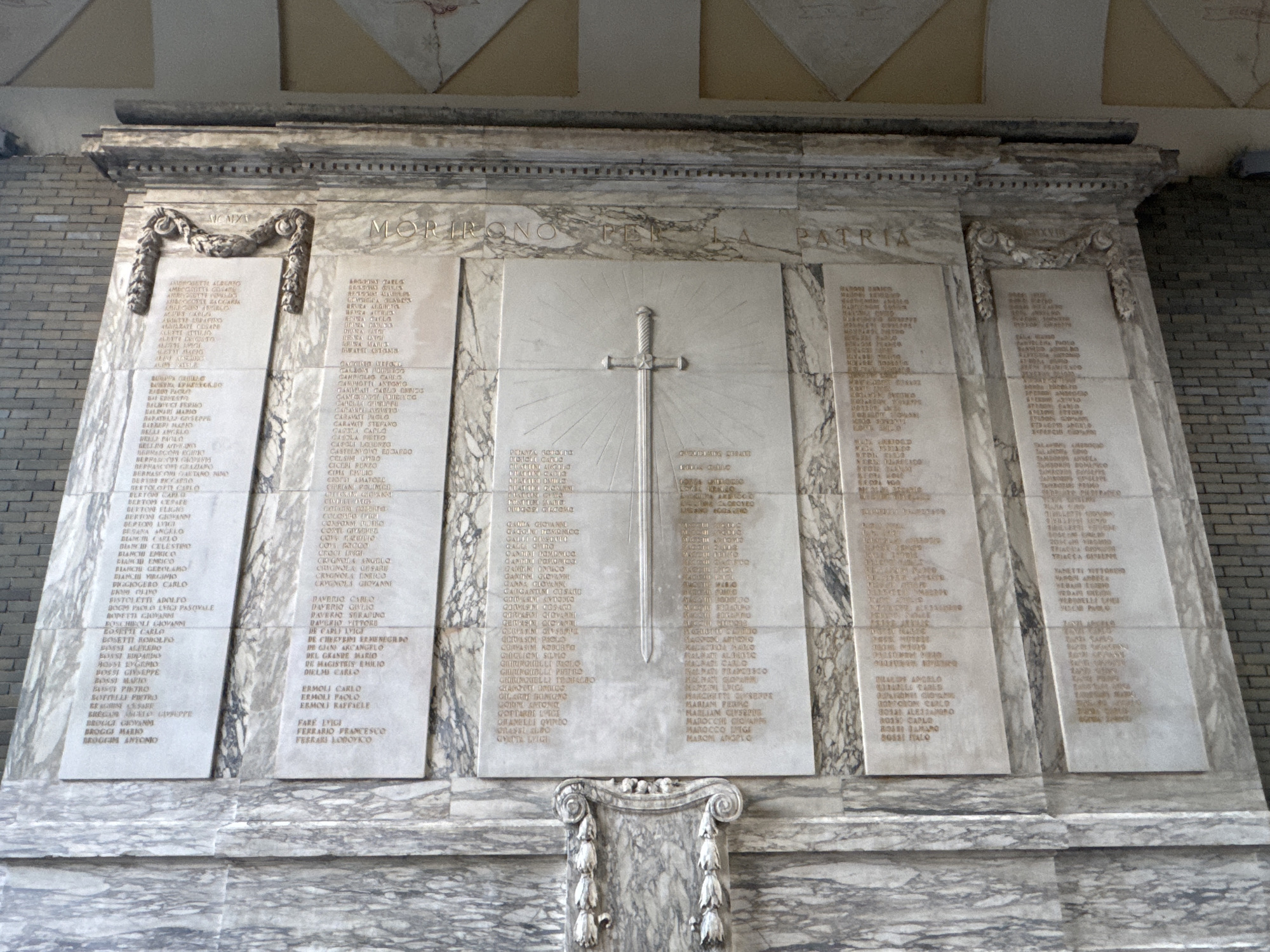 The image shows a marble monument with multiple columns of inscribed names. The monument is located in Varese, Italy, and is likely a war memorial. The monument is inscribed with the phrase "MORIRONO PER LA PATRIA," which translates to "They Died for the Fatherland." A prominent sword symbol is etched into the center panel, with rays emanating from it. The monument is decorated with ornate carvings and is set against a brick wall. The overall scene is solemn and commemorative.