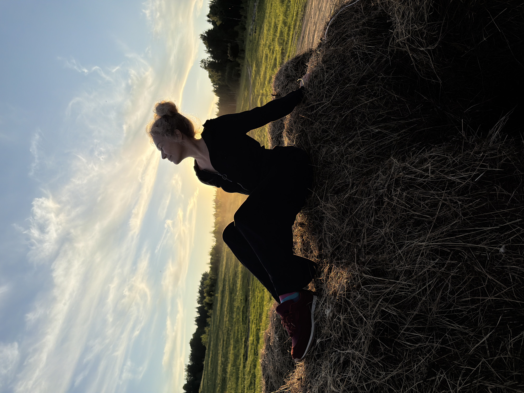 An outdoor shot captured during the golden hour, likely at sunset or sunrise, depicts a person silhouetted against a bright, cloudy sky and a sun-drenched field. The image is oriented with the horizon running diagonally from the top left to the bottom right.
The central subject is a person, appearing female, with hair styled in a bun. She is in a crouched or squatting position, leaning forward and resting her hands on a large, dark hay bale or stack of dried grass that occupies the right side of the frame. She wears dark, long-sleeved clothing and dark red sneakers with vibrant, multi-colored socks visible at the ankles. Her profile is visible, with her head tilted downwards. The bright light from behind illuminates the edges of her hair and body, creating a strong silhouette.
In the background, a vast, green grassy field extends towards a dark treeline under a bright sky. The sky is predominantly light blue with soft, wispy clouds stretching across it, glowing with warm yellow and orange hues near the horizon, indicating the low position of the sun. The scene suggests a peaceful, rural setting, potentially in the Аист Снт area of Russia, characterized by open fields and distant forest. No visible text is present.