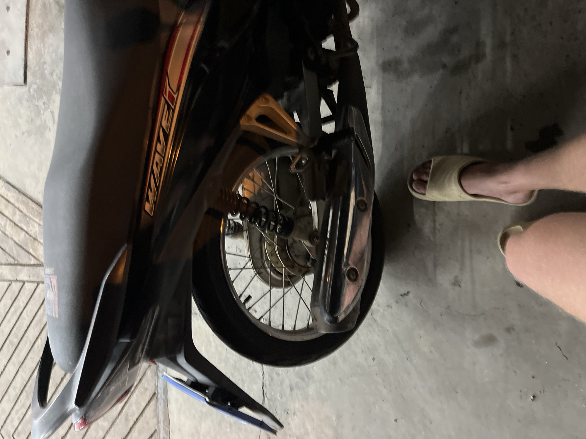 The image shows the rear portion of a black Honda Wave scooter parked on a concrete floor. A person's legs and feet, wearing light-colored slippers, are visible in the foreground. The scooter's seat, taillight, rear wheel, exhaust pipe, and part of the "WAVE i" logo are visible. The background consists of a textured wall. The scene is outdoors, likely in a parking area or street in Chalong, Thailand, suggested by the scooter model and common setting.