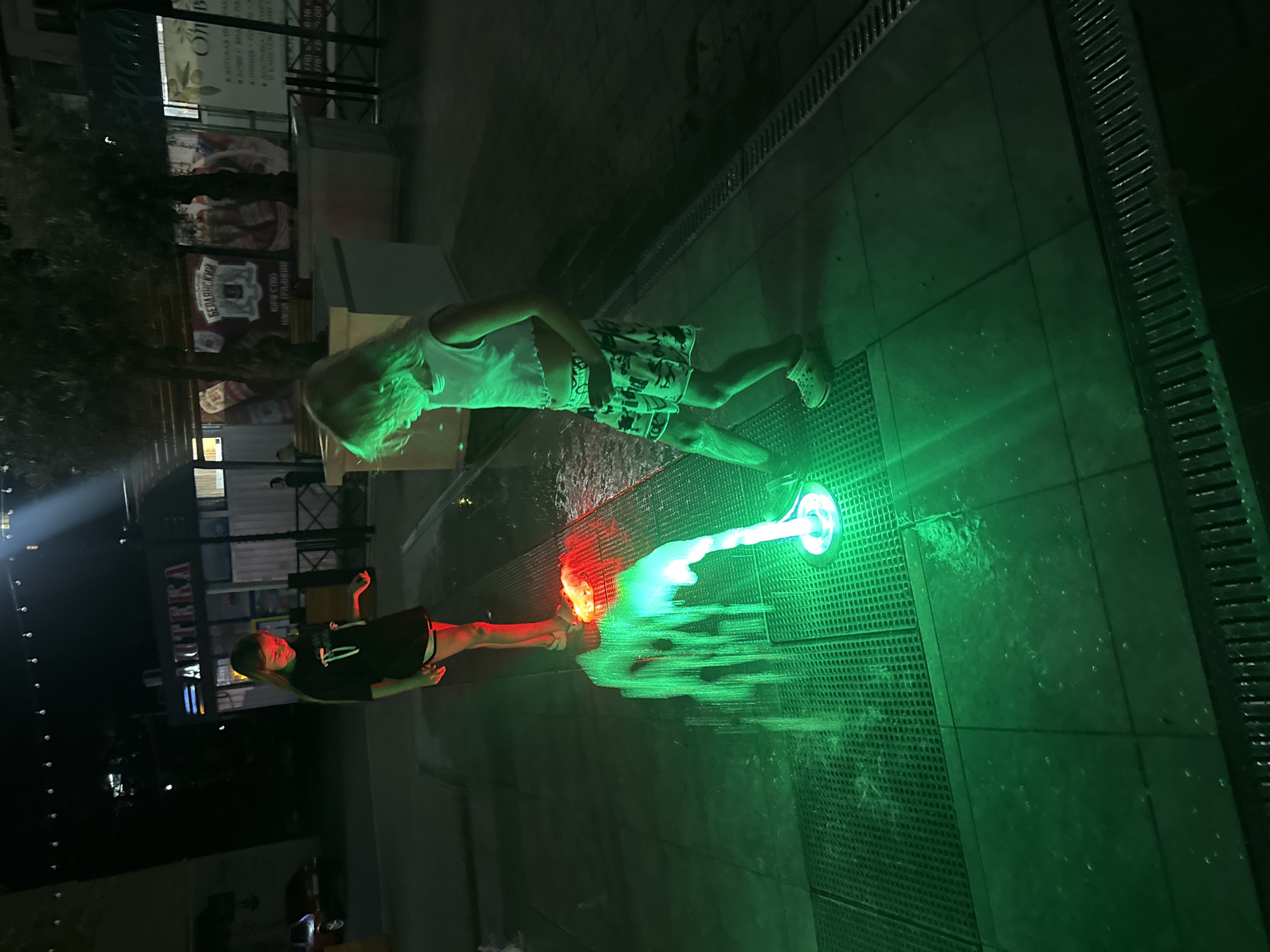 At night in Partenit, Ukraine, two young girls are playing in a public square with a fountain. The fountain has lights that change color, illuminating the water in green and red. The older girl, on the left, wears a black t-shirt and white shorts, and is looking down at the water. The younger girl, on the right, wears a white crop top and a patterned skirt, and is looking to her left. The square is paved with dark tiles, and there are planters with trees visible in the background. The buildings in the background are illuminated by lights, and there are strings of lights overhead. The scene appears to be a recreational area, possibly a promenade or park, and the presence of the fountain and the lighting suggest it is a popular spot for evening activities.