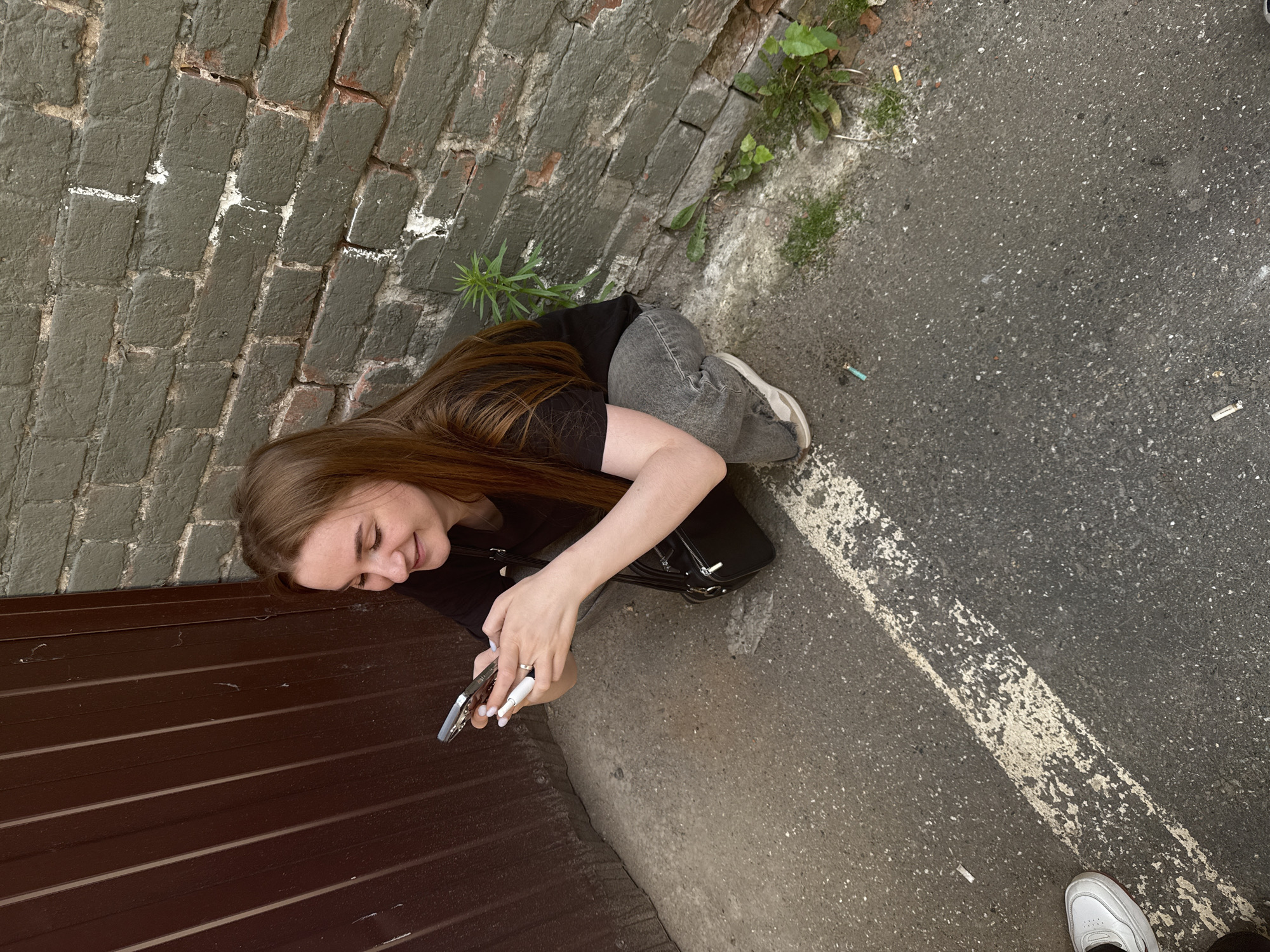 A young woman with long, light brown hair is crouched on an asphalt surface next to a brick wall and a dark brown corrugated metal surface. She is wearing a black t-shirt, grey jeans, and light-colored sneakers. She is holding a smartphone in her left hand and a white object, possibly an e-cigarette, in her right hand. A black shoulder bag is behind her. The brick wall is painted a dark grey with some white paint splatters. Several small plants are growing in the cracks of the wall and near its base. The asphalt surface has a faded white line painted across it. There are cigarette butts scattered on the ground. The scene appears to be an outdoor urban environment, potentially an alleyway or service entrance. The location is identified as the city of Perm, Russia.