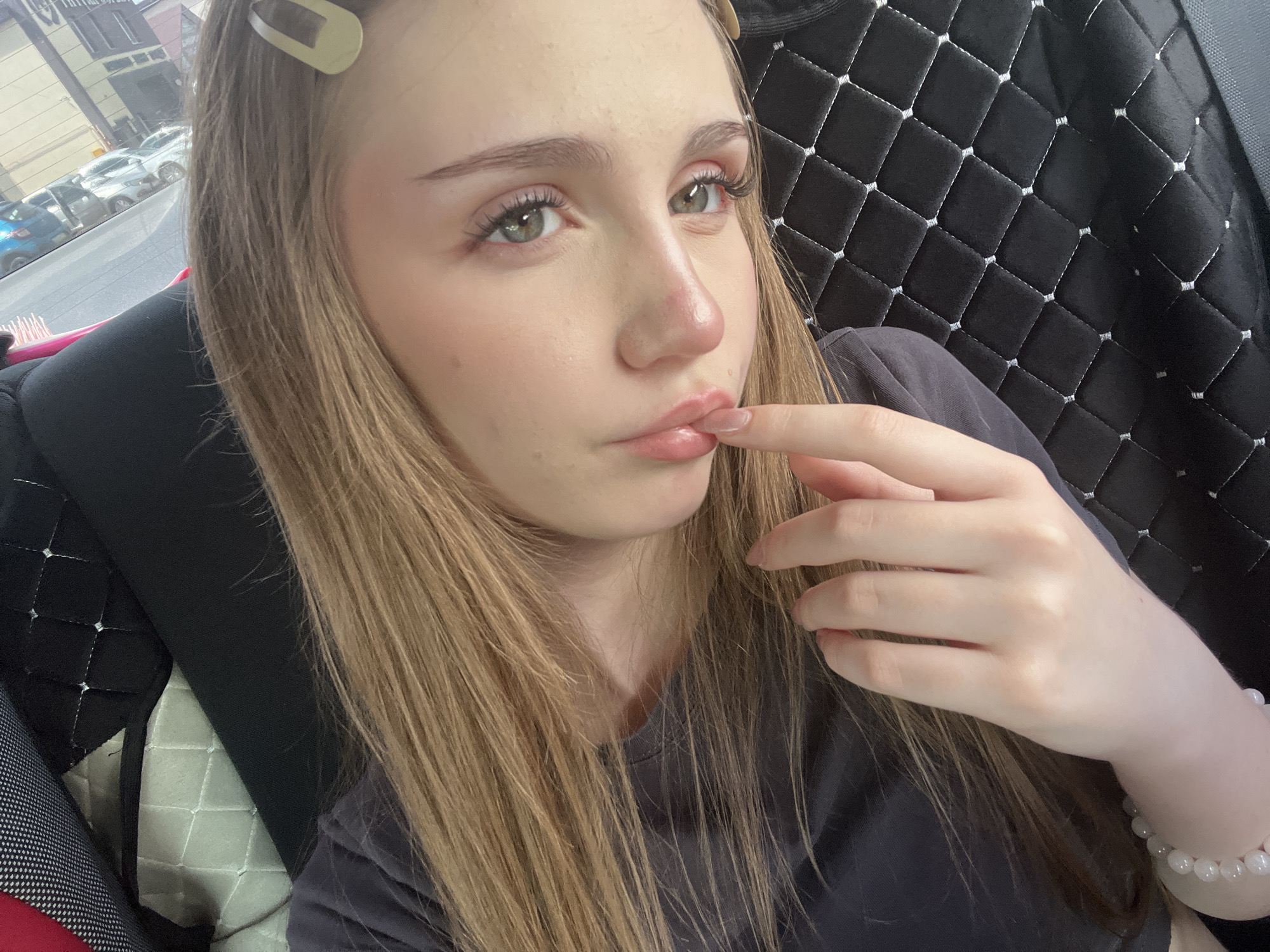 A young woman with long, light brown hair, a light complexion, and green eyes is captured in a close-up, selfie-style photograph. She is wearing a dark gray t-shirt and a bracelet of round, white beads on her left wrist. Her right hand is brought up to her lips, with her index finger touching her lower lip. She has a subtle makeup application, with noticeable mascara and blush. A beige hair clip is visible in her hair near the top of her head. The background suggests she is inside a vehicle, with a quilted black seat cover with white stitching visible behind her to the right. To the left of the frame, out of focus, is a street scene with several parked cars, suggesting an urban environment. The lighting indicates it is daytime, with natural light illuminating her face. The image is likely taken in the city of Novaya Otradovka, Russia, given the context of the request.