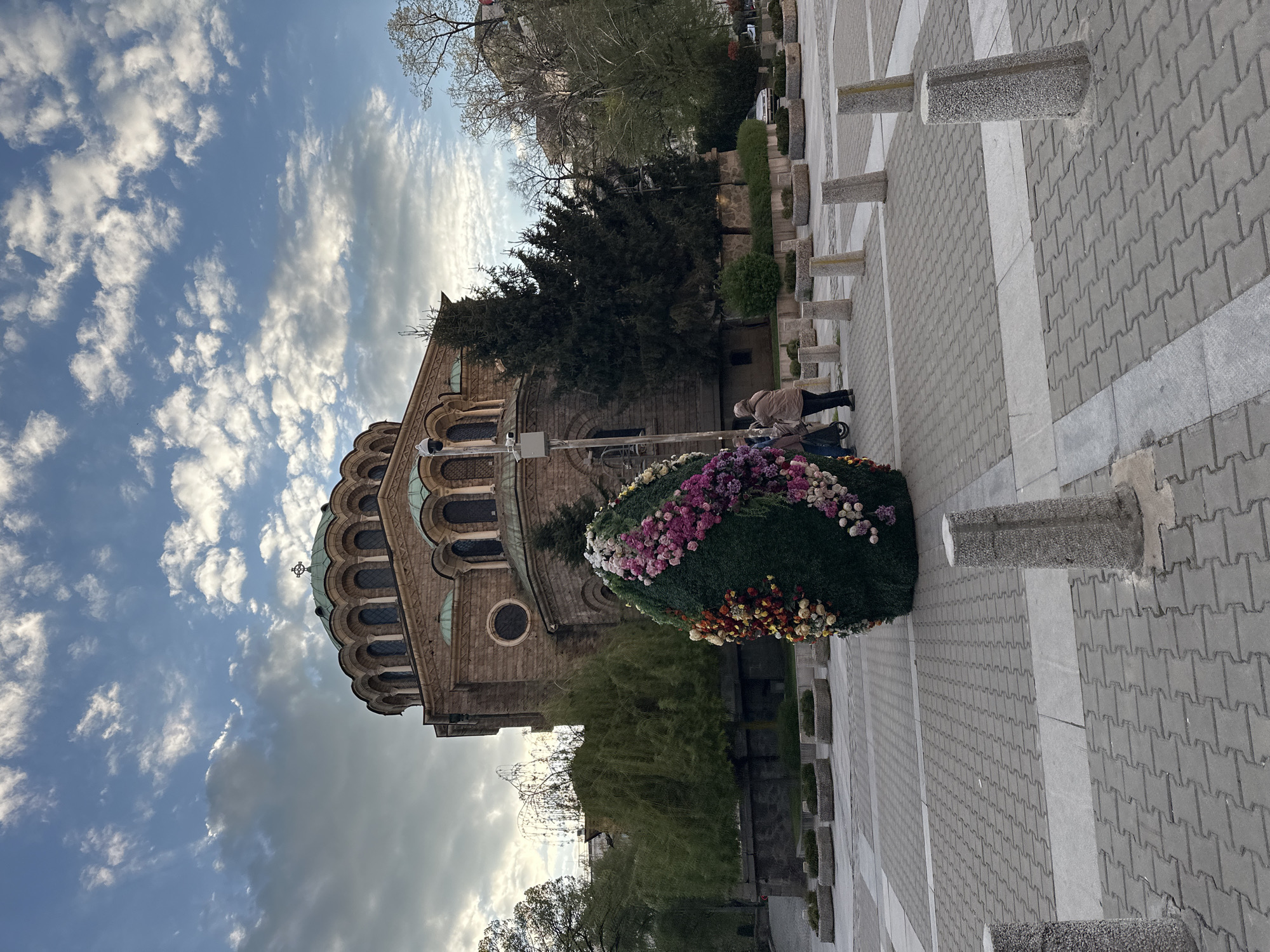 The image displays an outdoor scene featuring a large Eastern Orthodox church or cathedral, identifiable by its distinctive domes, arched windows, and stone construction. In the foreground, a prominent oval-shaped floral display, lush with green foliage and a vibrant array of pink, purple, yellow, white, orange, and red flowers, draws attention.
A lone individual, dressed in a light-colored jacket, stands near this floral arrangement, appearing to observe it. The ground consists of light grey interlocking pavers interspersed with smoother concrete sections. A series of grey stone bollards line the paved area, separating it from a landscaped section with various trees and bushes, including a large evergreen conifer. The church's architecture suggests a historical and significant religious site.
The sky is partially cloudy, with clear blue patches indicating a daytime setting, likely during pleasant weather. No specific event is explicitly shown, but the elaborate floral decoration implies a celebration, religious holiday, or public commemoration. The scene takes place in what appears to be a public square or churchyard, consistent with the provided context of Sofia, Bulgaria. There is no visible text in the image.