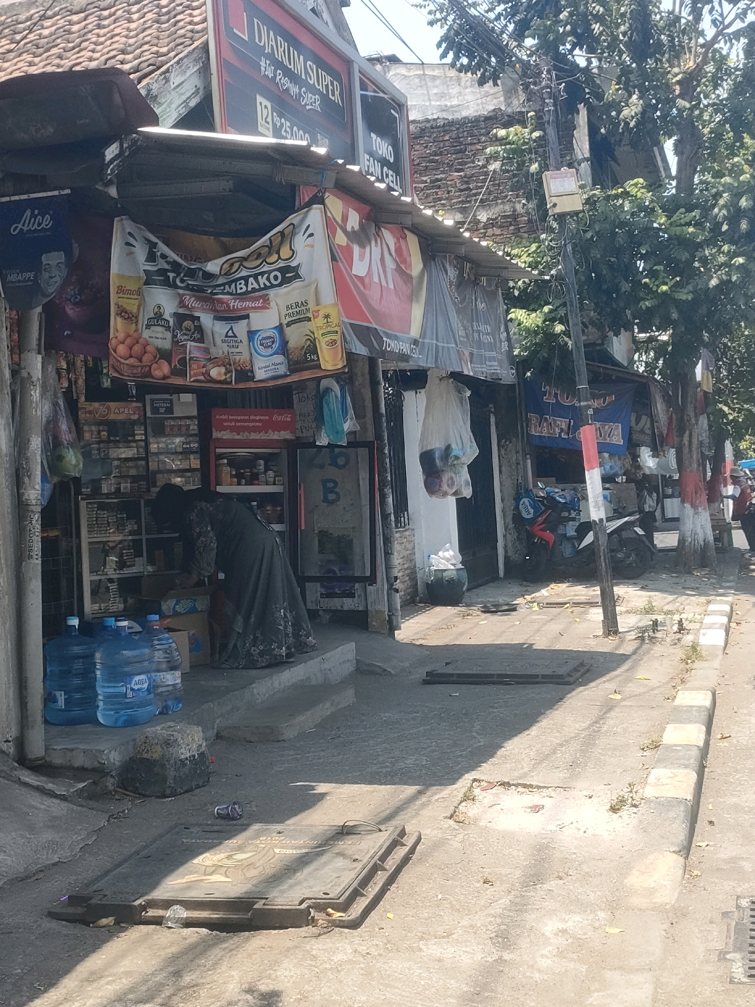 The image shows a street scene in Surabaya, Indonesia, in front of a small shop. The shop has a red and white striped pole in front of it and a banner advertising "TOKO RAFIFA". A person in a dark dress is standing in front of the shop, looking down at shelves stocked with goods. In front of the shop, there are several large blue water jugs. The street is paved and there are some manhole covers in the foreground. Trees and buildings are visible in the background. The weather appears to be sunny.
