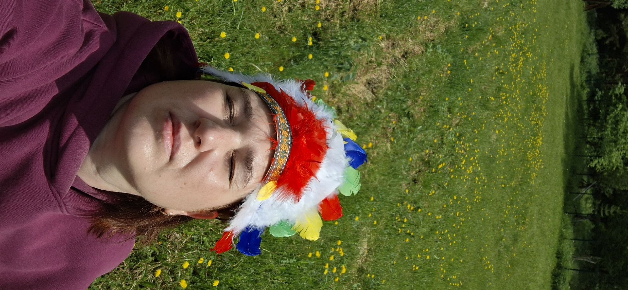 A person with reddish-brown hair lies on a grassy lawn dotted with yellow dandelions, wearing a burgundy hooded sweatshirt and a colorful feathered headdress reminiscent of Native American styles. The scene is bathed in sunlight, casting shadows on the person's face and clothing. The background features a field of dandelions and a line of trees along the edge of the lawn. The exact location cannot be determined with the given information, except for the stated city of Прикамский, Russia.