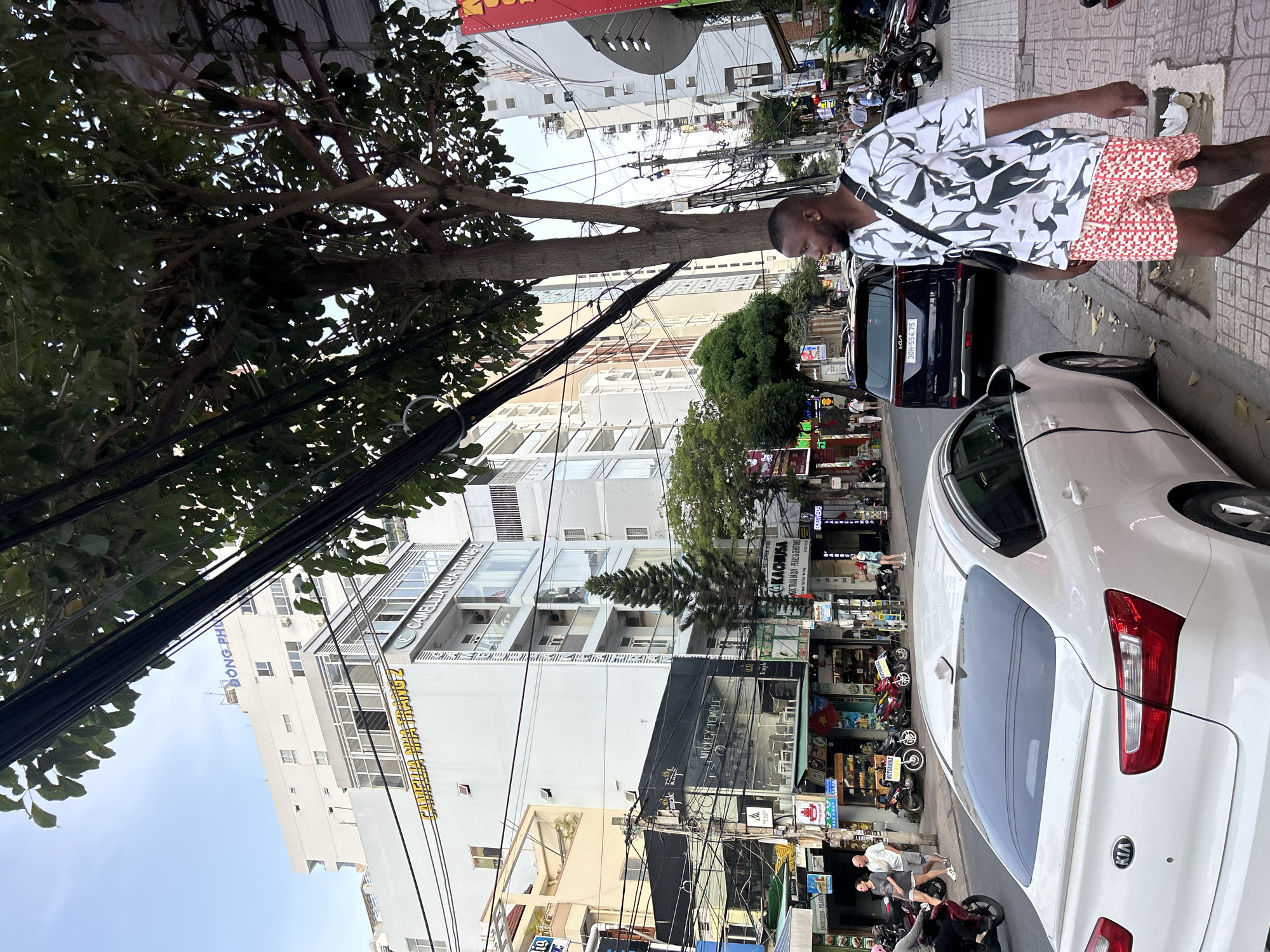 A Black man wearing a white shirt with a black abstract pattern and red and white checkered shorts walks on a sidewalk in Nha Trang Ward, Vietnam. He has a black crossbody bag slung over his right shoulder. In the background, a white Kia sedan is parked, and a dark blue SUV is driving down the street. The street is lined with buildings, many of which have balconies and signs in Vietnamese. A large tree with dark green leaves is prominent on the right side of the image, casting shadows on the sidewalk. Power lines are visible crisscrossing overhead. Several motorbikes are parked on the side of the street, and a few people are walking in the distance. The sky is bright and appears to be a clear day.