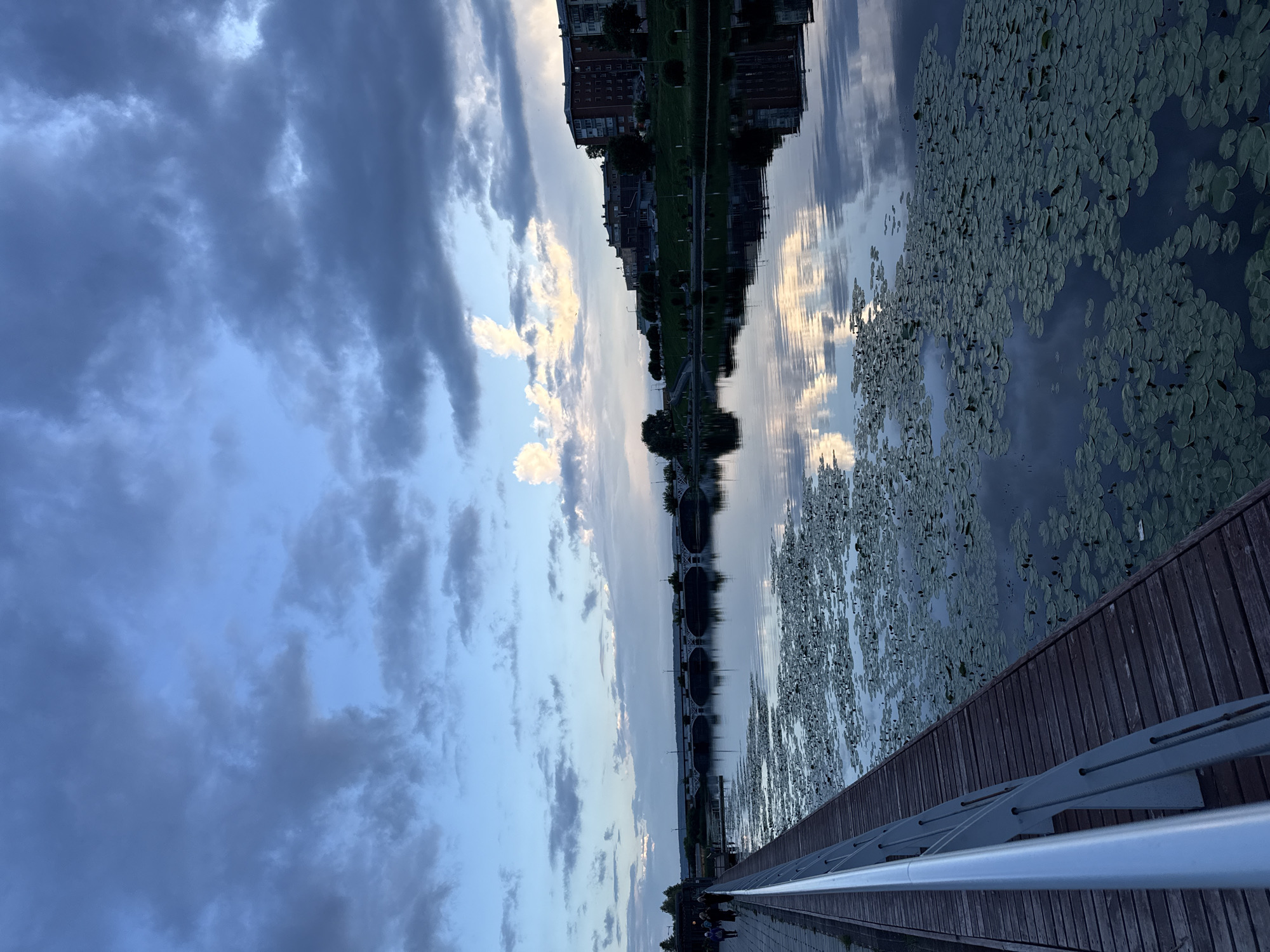 This is a vertical, eye-level, outdoor photograph taken at dusk on a riverfront in Votkinsk, Russia. The foreground features a wooden boardwalk with a modern silver railing. Several people are walking along the boardwalk on the left side of the frame, appearing as silhouettes. The river occupies the majority of the image, with the water covered in numerous lily pads. The surface of the river reflects the cloudy sky and the buildings on the opposite bank.
In the middle ground, a long, arched stone bridge spans the river. On the far bank, a grassy slope leads up to a row of multi-story brick buildings and trees, silhouetted against the twilight sky. The sky is filled with dramatic clouds, a mix of dark grays and lighter blues, with hints of illuminated clouds in the distance. The overall mood is serene and contemplative, characteristic of the evening hours.