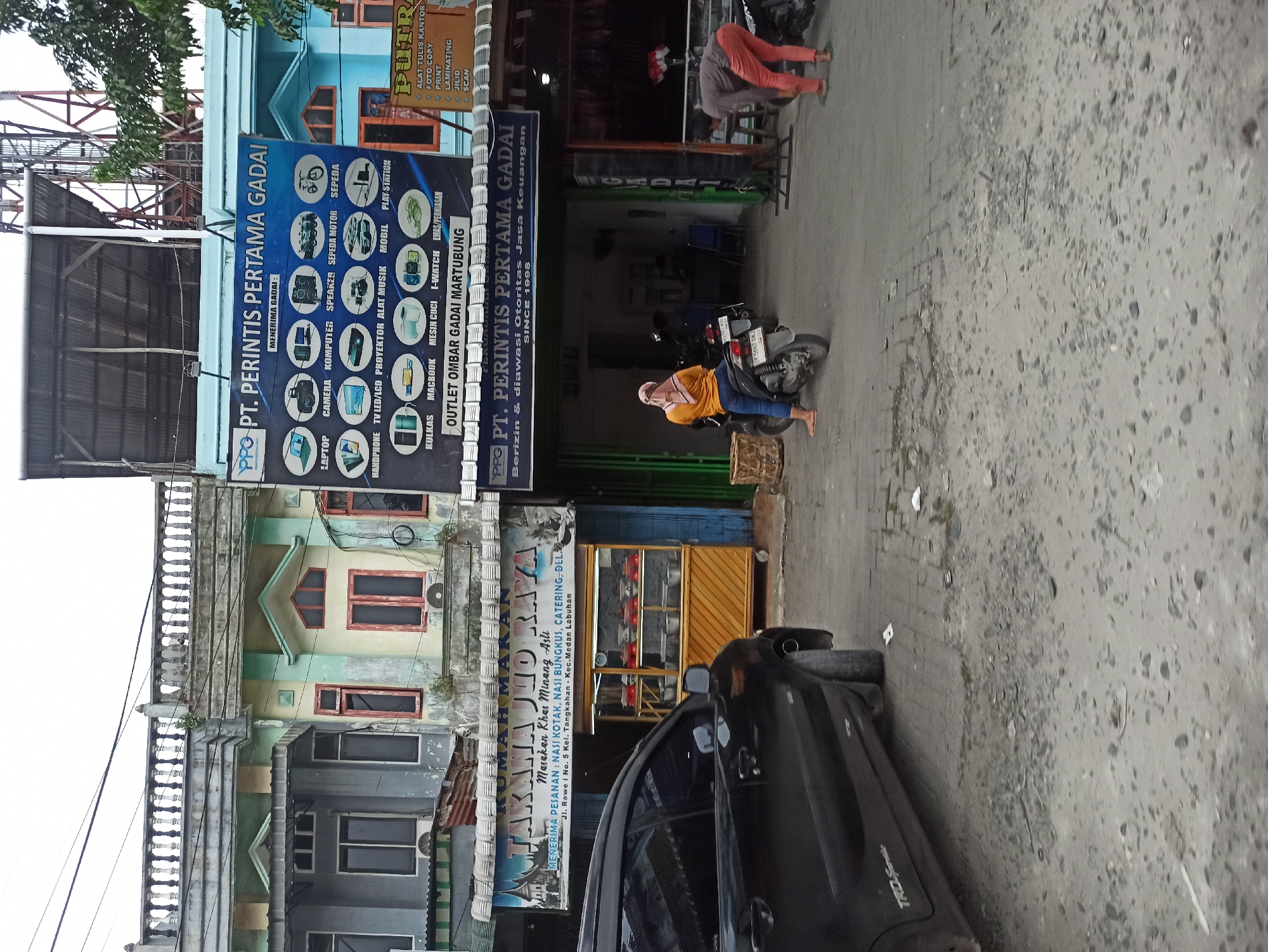 The image captures a street scene in Medan, Indonesia, featuring several establishments. On the left, a black car is parked, with part of its rear and side visible. Adjacent to the car is a food stall labeled "RUMAH MAKAN TAKANA JILO RAYA" with a display of food items in glass containers. To the right of the food stall, a large banner for "PT. PERINTIS PERTAMA GADAI" advertises various services including electronics, vehicles, and household appliances. Another establishment labeled "PUTRA ALAT TULIS KANTOR" appears to be an office supply store. In the foreground, a woman wearing a headscarf and an orange top sits on a motorcycle. Further to the right, a person in orange pants is attending to something on the ground, possibly near a motorcycle. The street is paved with bricks and has areas of loose gravel and debris. The buildings exhibit a mix of architectural styles and ages, with some showing signs of wear and tear. Overhead, power lines are visible against a cloudy sky.