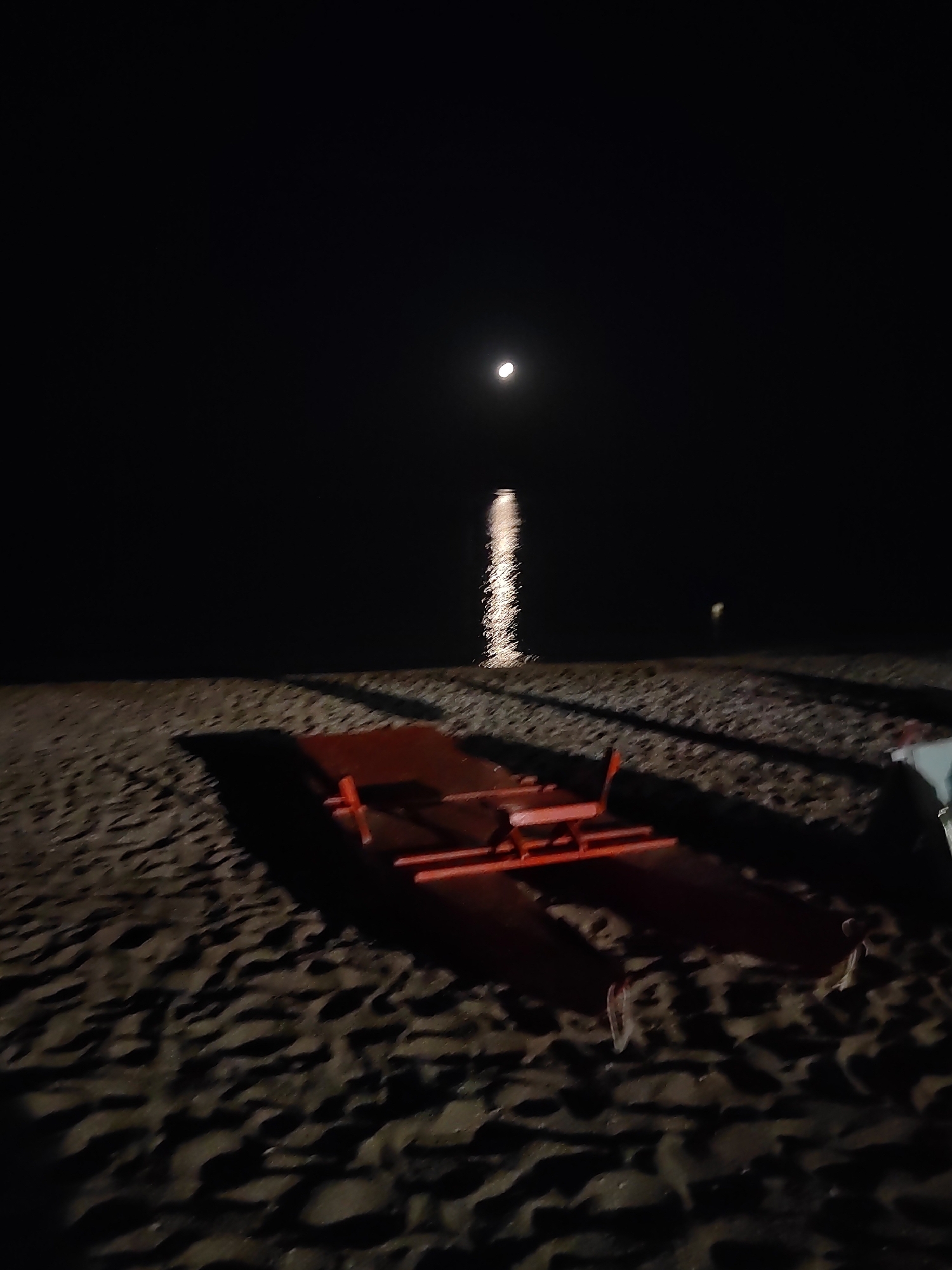 This is a nighttime photograph taken on the beach in Porto Recanati, Italy. The scene is illuminated by the moon, which appears as a bright orb in the dark sky, casting a shimmering reflection on the water. A red lifeguard rescue boat is positioned on the sand in the foreground, with its shadow stretching across the rippled sand. The beach is dark and appears to be largely empty, with the exception of some indistinct shapes in the distance and possibly a small, distant light source on the water. The overall atmosphere is serene and somewhat mysterious due to the low light conditions.