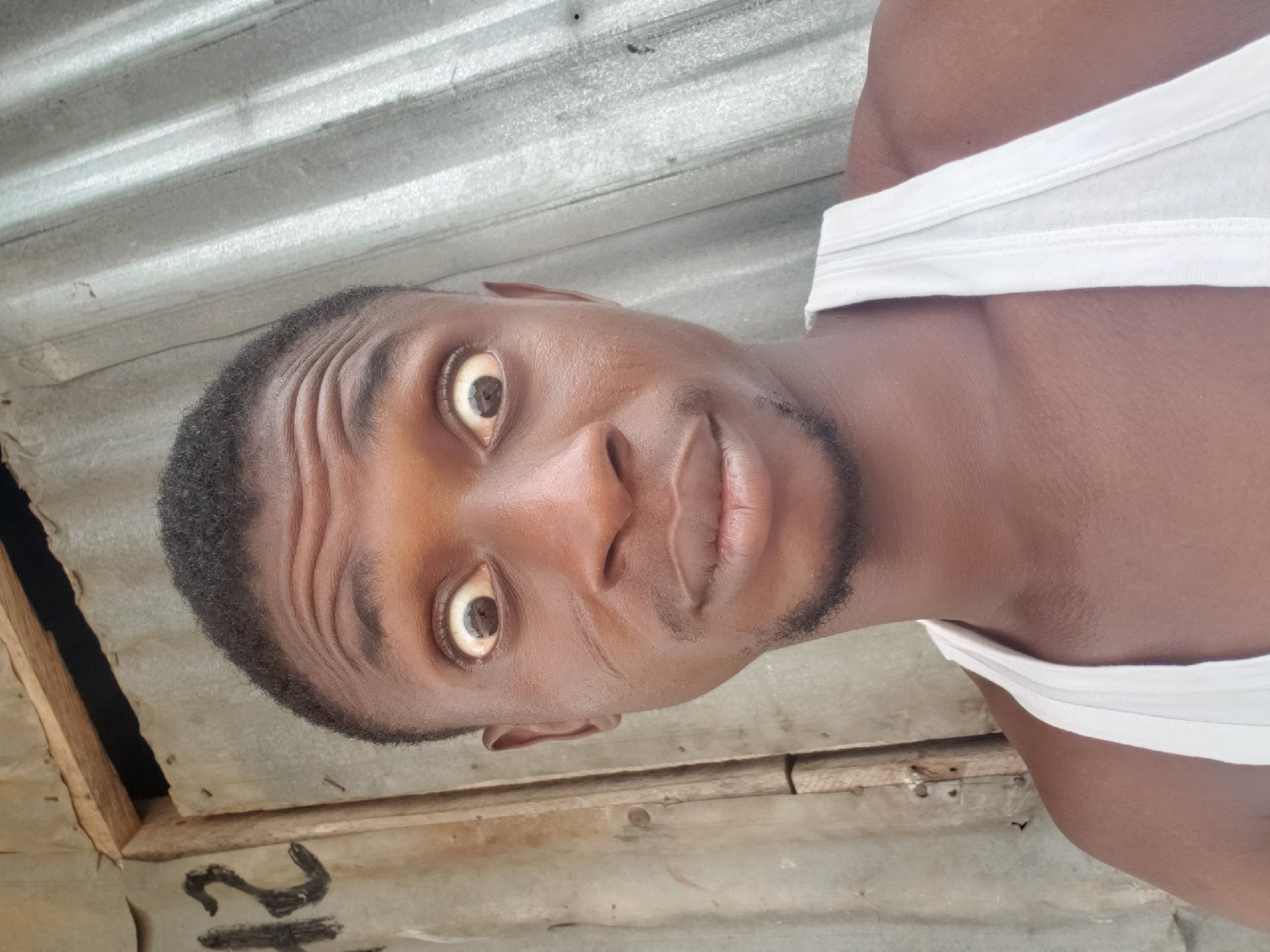The image is a close-up portrait of a young man with dark skin, shown in a horizontal orientation. He gazes directly at the camera with wide, prominent eyes, exhibiting an expression of surprise or intensity. His eyebrows are raised, causing several horizontal wrinkles to form across his forehead. He has short, dark, curly hair and a faint beard or stubble outlining his jaw and chin. His lips are slightly parted. The man is wearing a simple white sleeveless undershirt or tank top, visible across his shoulder and upper chest.
The background features corrugated metal sheeting, likely a wall or roof, with distinct parallel ridges and a weathered, grayish-white texture. In the bottom left section of the image (when oriented upright), wooden structural elements, possibly beams or framing, are visible. Beneath these wooden pieces, faint black markings are present on the corrugated surface, resembling the number "2" and part of another character. The lighting is bright and even, suggesting it was taken during the day. The setting appears informal or rustic, aligning with the provided location context of Adankolo, Nigeria.