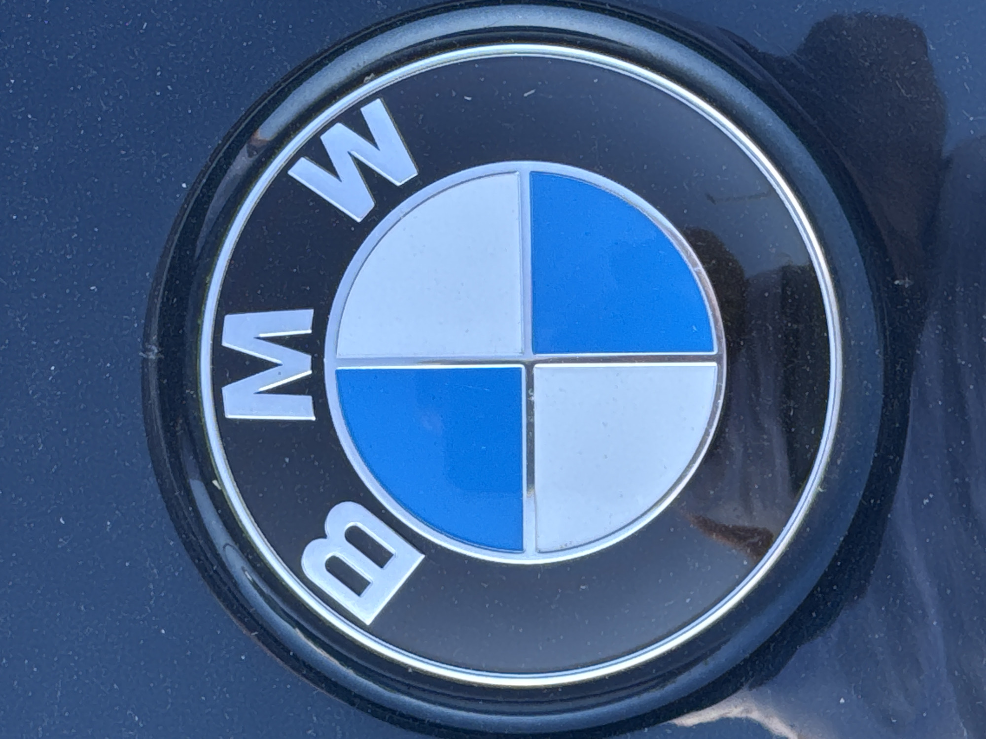 The image shows a close-up of a BMW emblem. The emblem is circular and features the iconic blue and white quadrants. The letters "B", "M", and "W" are visible in silver around the circular emblem, arranged in a clockwise direction. The emblem appears to be affixed to a dark, possibly black, surface which is dusty or shows signs of wear. The lighting suggests an outdoor environment, and the overall appearance is that of a car badge. The image does not provide any specific information about the city of Wettstetten, Germany, or any people or actions occurring in the scene.