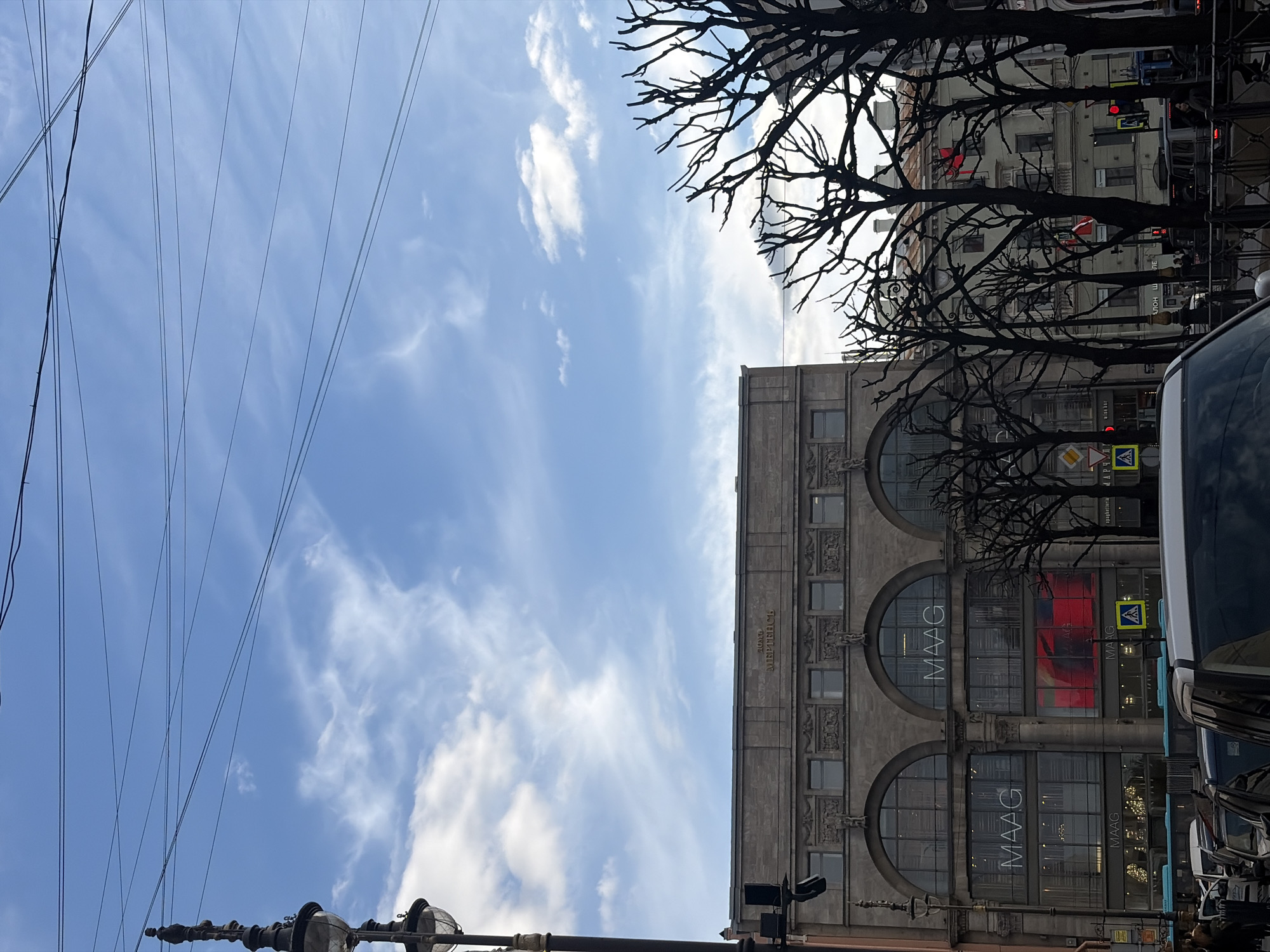 The image shows a street scene in Saint Petersburg, Russia. The dominant architectural feature is a large building with arched windows, displaying the word "MAAG" on its facade. Above the building, electrical wires intersect the sky. Bare tree branches frame the upper right portion of the image. Traffic lights and pedestrian crossing signs are visible, indicating the presence of a street. Vehicles are parked or moving along the street, with a partial view of a car on the right. The sky is blue with scattered clouds.