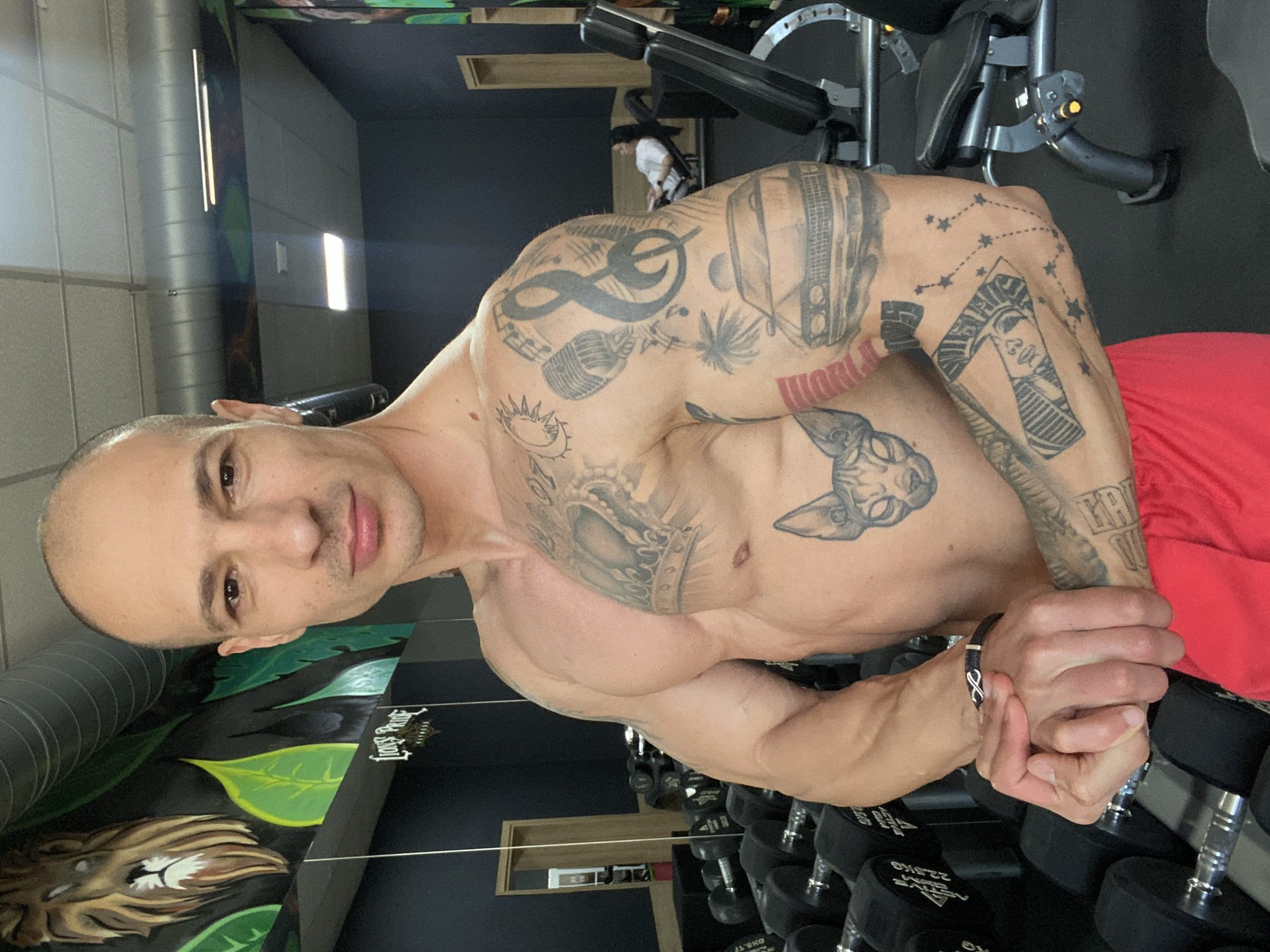 A bald, shirtless man with multiple tattoos on his arms and chest poses in a gym. He is wearing red shorts and a black bracelet. In the background are weight machines and dumbbells. A lion mural is visible on a wall. The location may be Pleven, Bulgaria, although this cannot be definitively confirmed from the image.