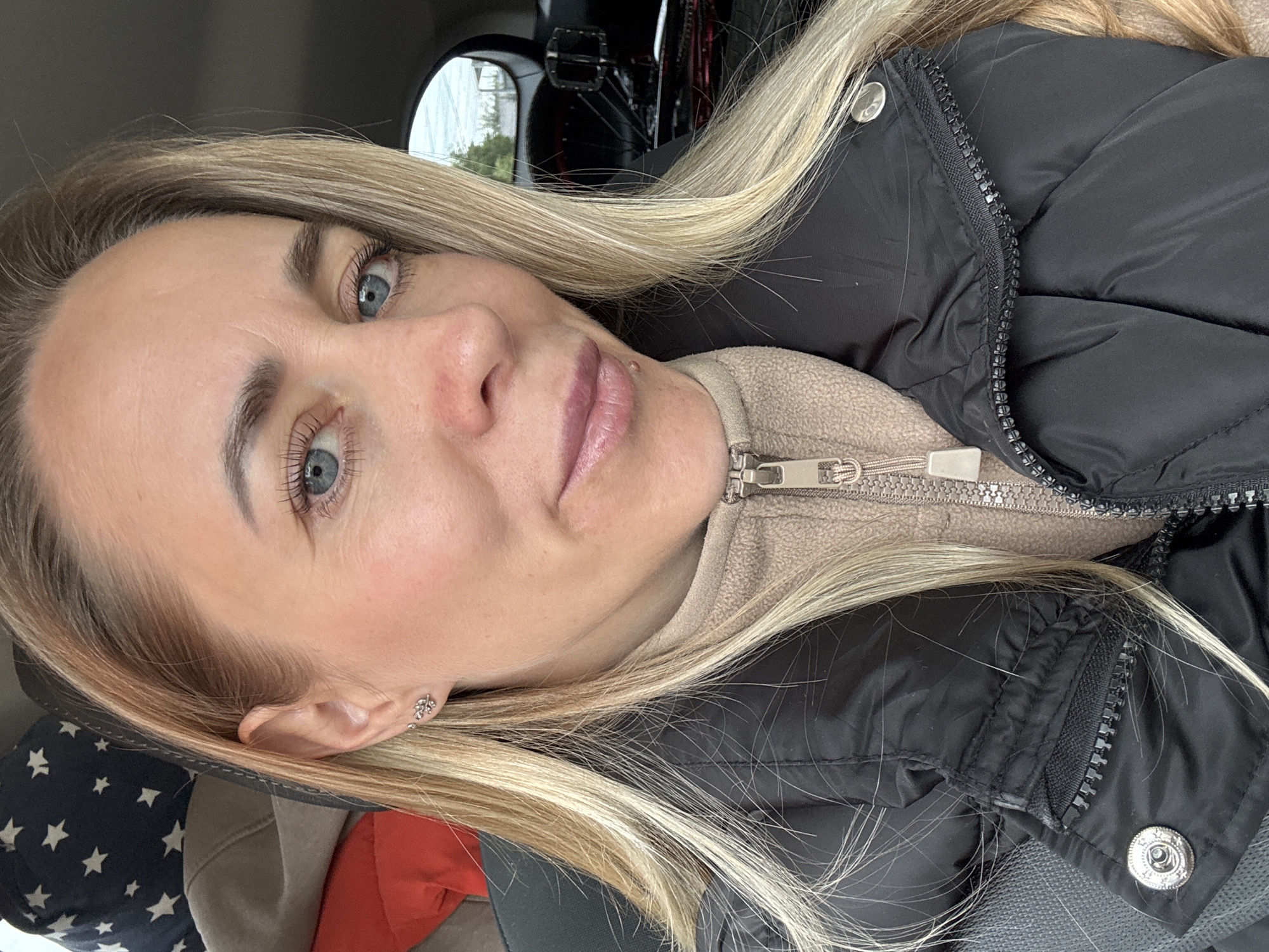 A close-up, tilted portrait shot captures a blonde woman with fair skin and light blue eyes. She is wearing a black puffer jacket over a beige fleece with a zipper. A silver stud earring is visible in her left ear. The background suggests she is inside a car, with a glimpse of the outside world visible through the rear window. There's a reflection visible in the rear mirror. It is impossible to determine the location in the city of Chaikovsky, Russia without further information from the image.