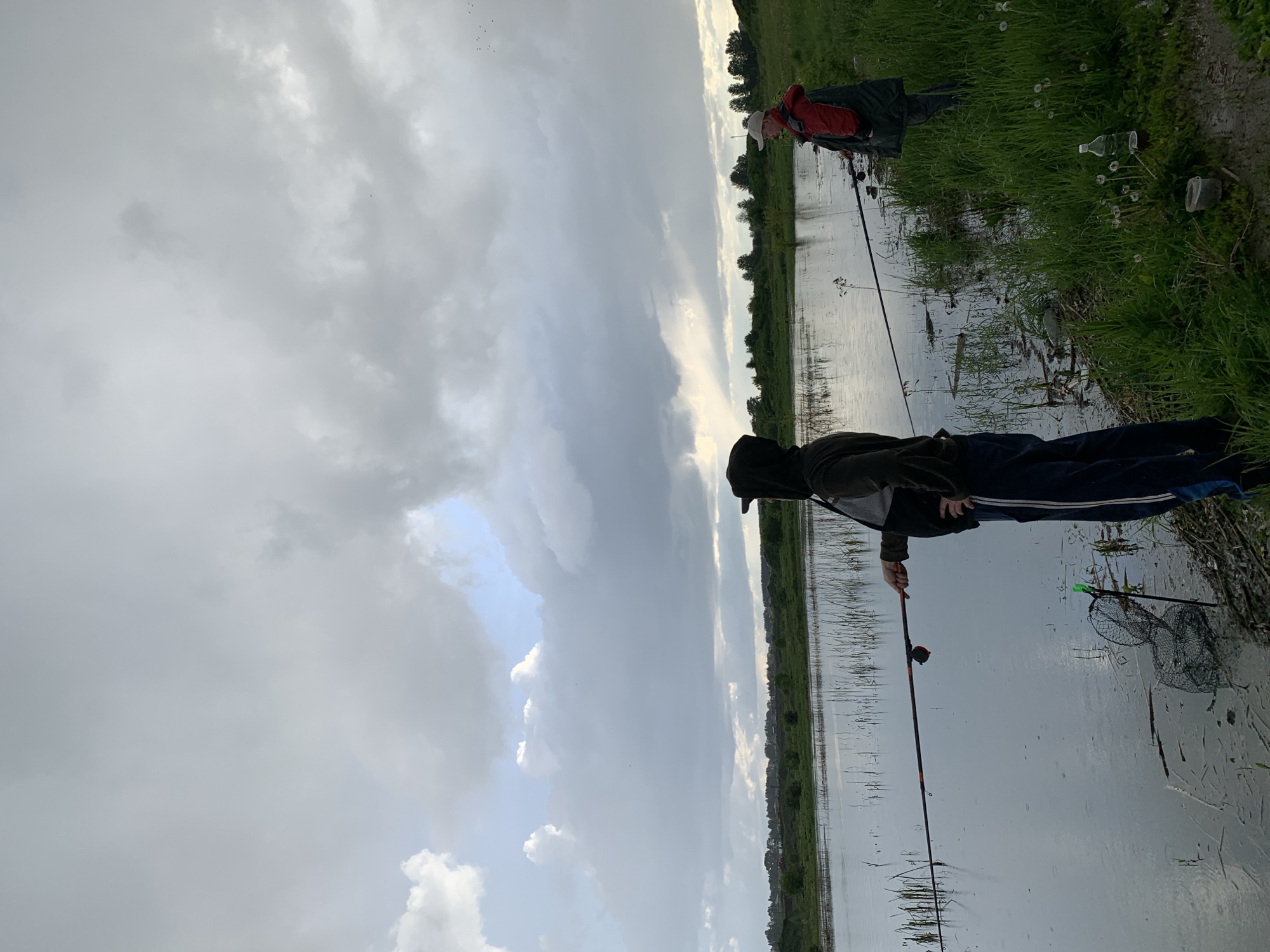 The photograph depicts two people fishing by a body of water under an overcast sky. The location appears to be near Shaberdino, Russia. In the foreground, a person in a hooded jacket and dark pants with a white stripe holds a fishing rod. A fishing net is partially submerged in the water near them. Another person, wearing a red jacket and a white cap, is positioned further away, also holding a fishing rod. The surrounding landscape consists of green grass and vegetation along the water's edge. Scattered trash, including plastic bottles and containers, is visible on the bank. The water reflects the cloudy sky, and reeds or aquatic plants are emerging from the water.