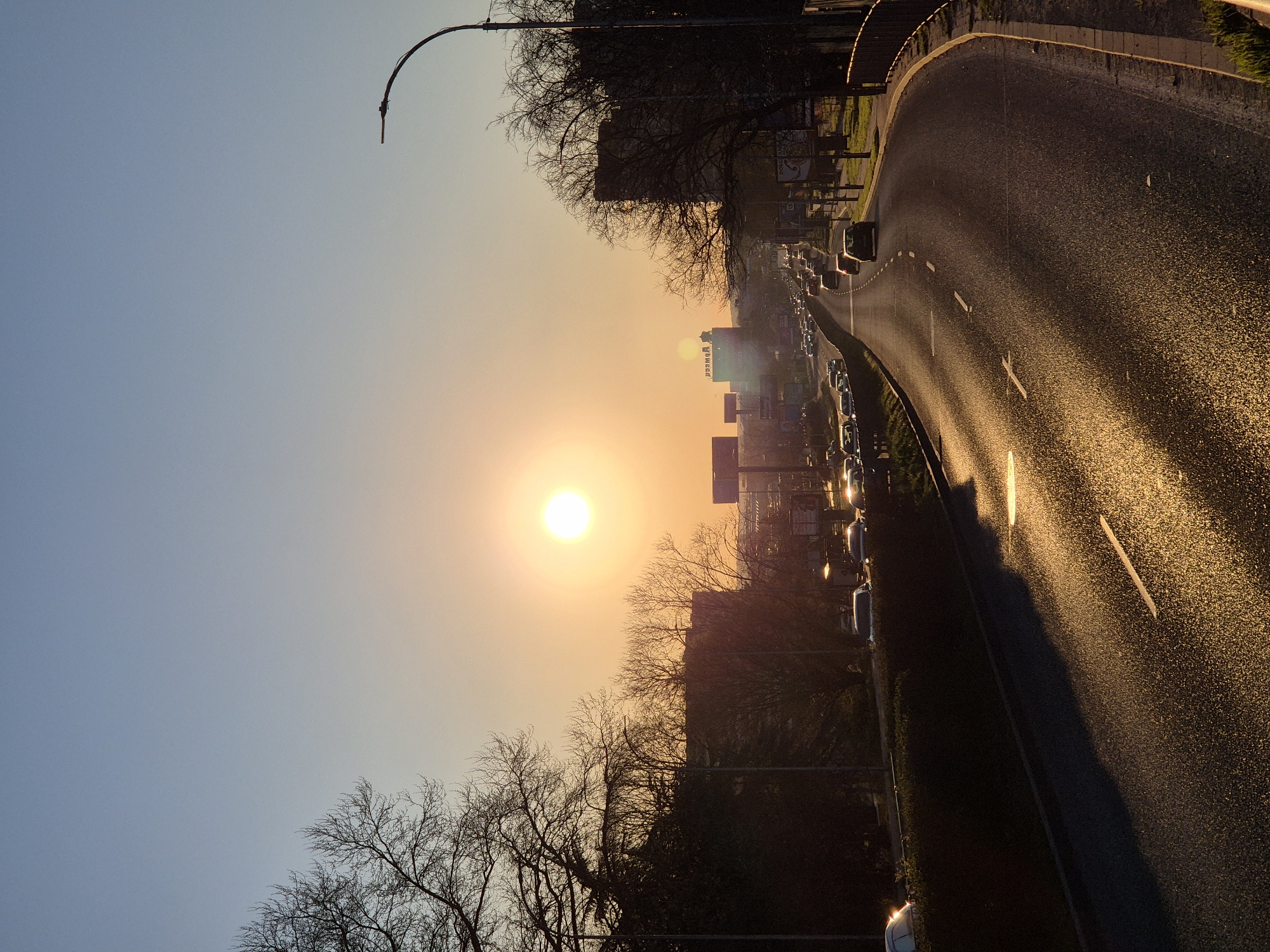 The image shows a road scene at sunset, possibly in an urban area, with the sun positioned centrally in the frame. Buildings and trees line the horizon, partially obscured by the sun's glare. Cars are visible on the road, and their headlights indicate they are facing the camera. The road surface reflects the golden light of the setting sun. A street light is visible on the left side of the road. The image is likely taken at sunset. It is impossible to say that this image is located in Varna, Bulgaria based solely on visual information.