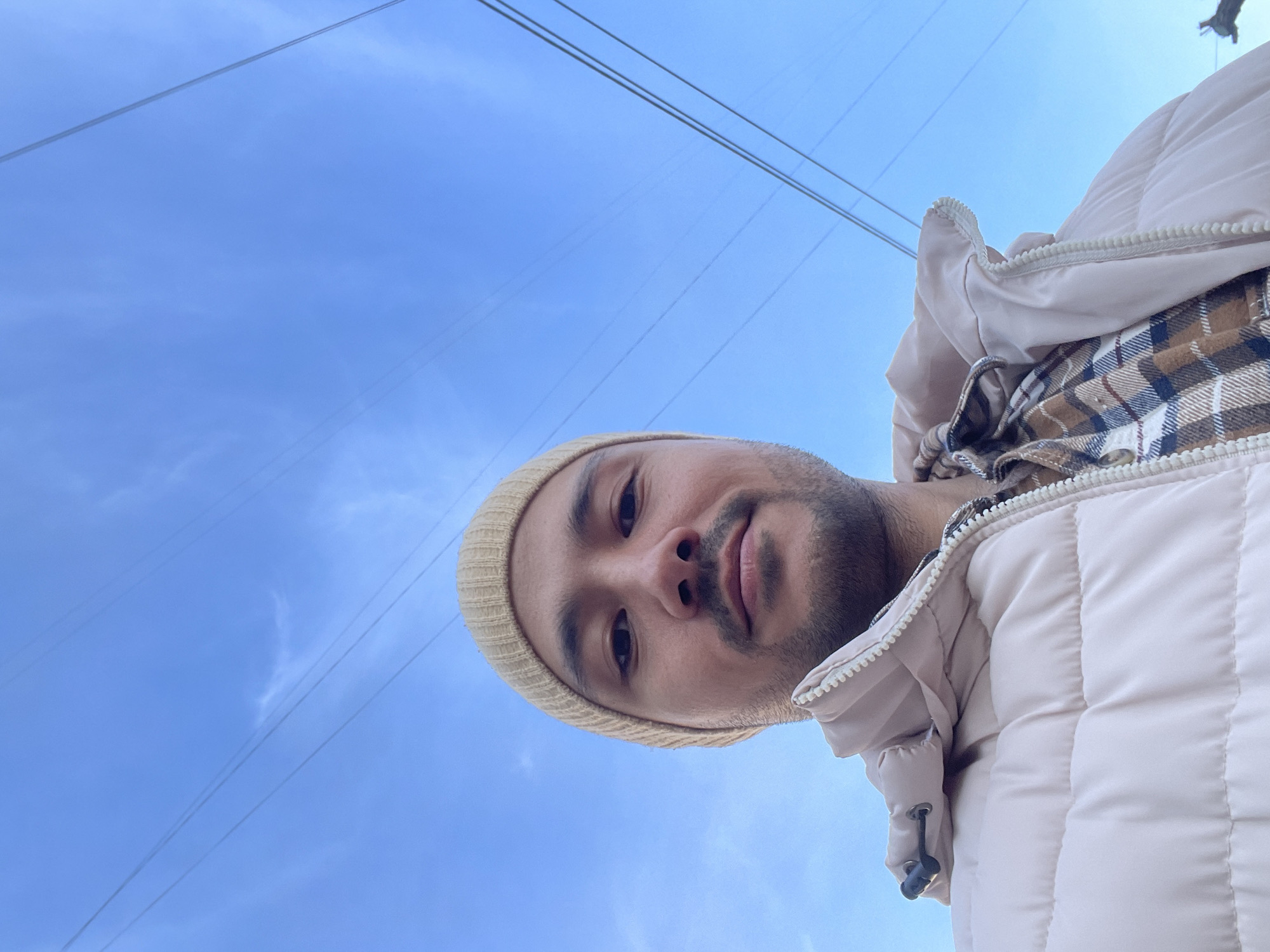 A selfie of a person with light brown skin, a beard, and wearing a tan beanie, a plaid shirt, and a light-colored puffer jacket is set against a light blue sky. Overhead power lines are visible in the sky. Without additional information, the location in Izhevsk, Russia, cannot be determined.