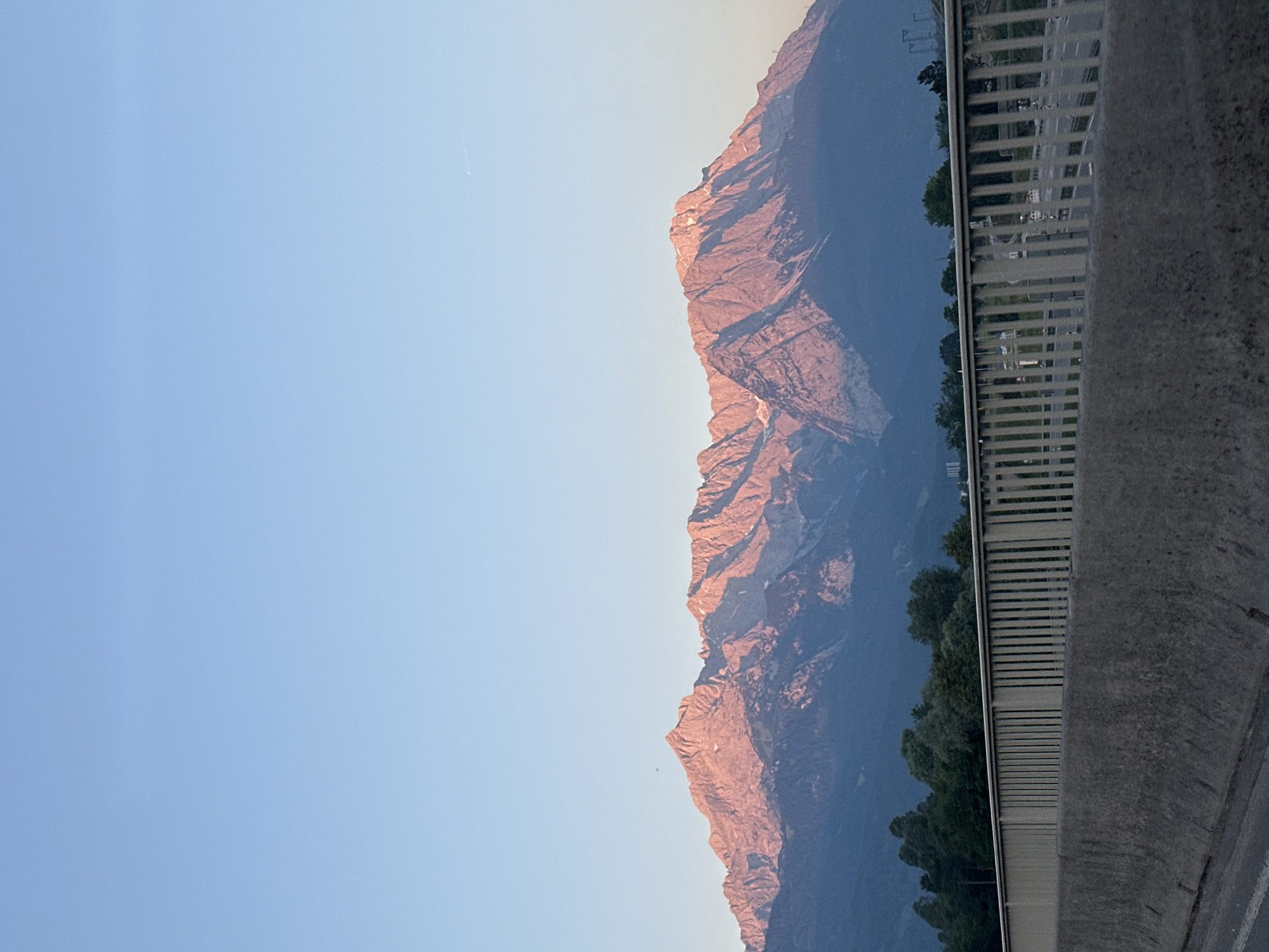 The image shows a panoramic view of the Alps mountains, bathed in the warm glow of sunset. In the foreground, a concrete barrier and a metal railing suggest the scene is captured from a road or bridge. The mountains, jagged and imposing, rise majestically against a clear, pale blue sky. Their peaks are illuminated by the setting sun, casting long shadows and highlighting the rugged terrain. A line of dark green trees forms a transition between the mountains and the foreground. In the distance, faint structures and utility poles can be seen, indicating the presence of civilization near the natural landscape. The overall impression is one of serene natural beauty, with a hint of human infrastructure.