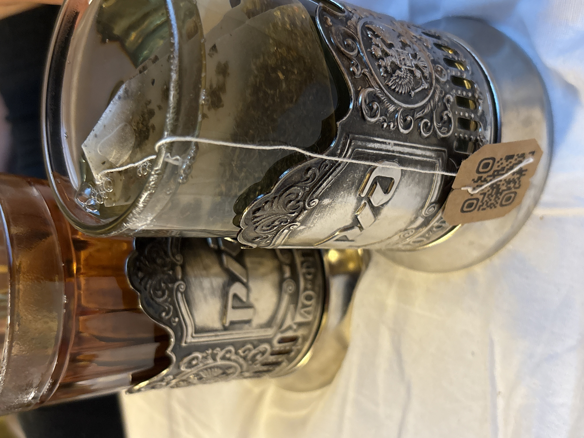 Two glasses of tea are shown in ornate silver holders on a white surface. The glass on the left contains amber-colored tea, while the glass on the right contains lighter-colored tea with a tea bag. Both holders have intricate embossed designs, including what appear to be Cyrillic letters and an emblem resembling the Russian coat of arms. A small brown tag with a QR code is attached to the holder of the glass on the right. The image is a close-up, focusing on the tea glasses and their holders. The background is out of focus, but appears to be a table covered with a white cloth. The scene suggests a moment of refreshment, possibly in a cafe or home setting within Khimki, Russia, given the cultural context of the tea holders and potential Russian emblems.