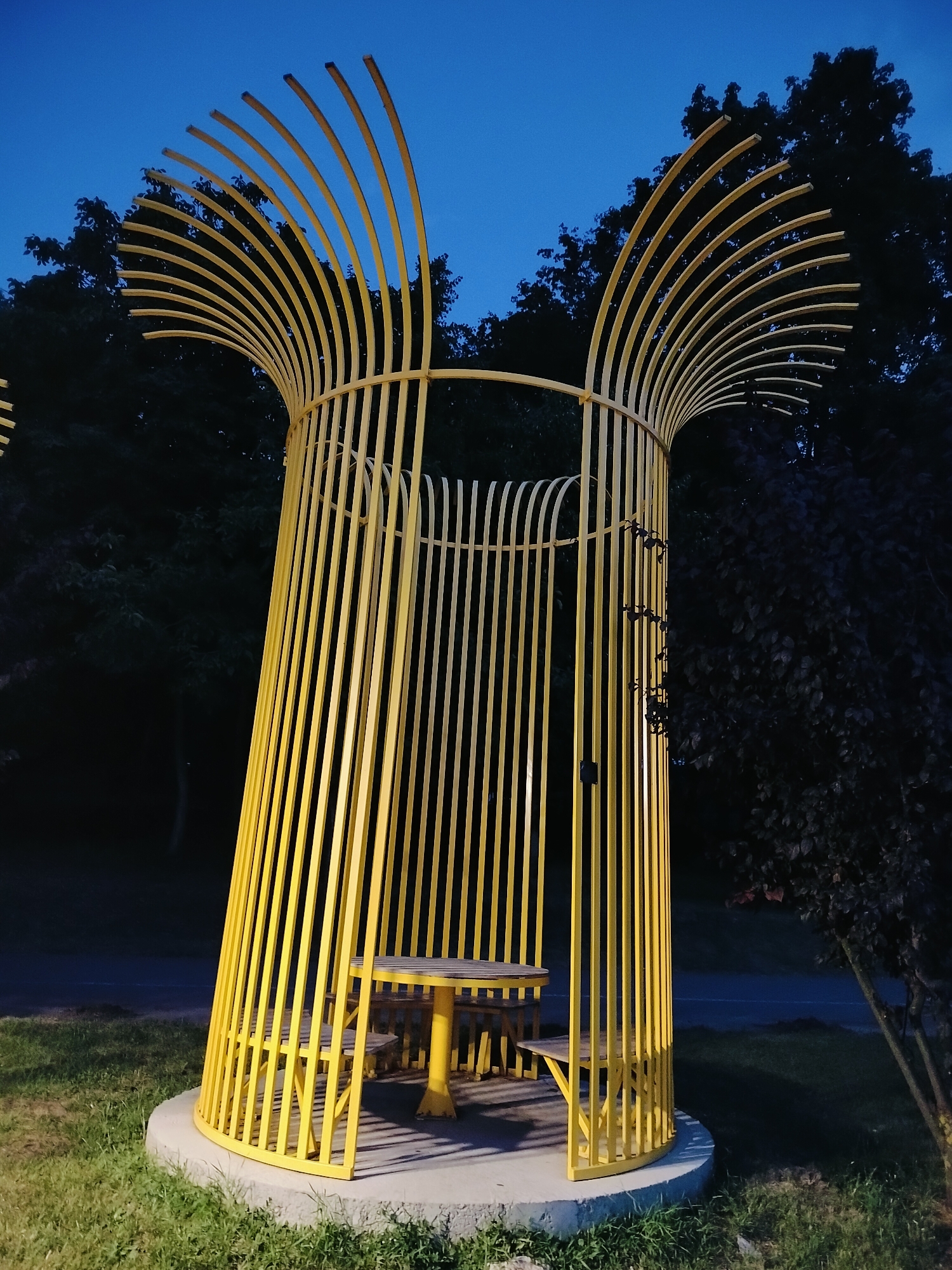 A bright yellow, cage-like gazebo stands in a park at dusk. The gazebo has a circular base and is constructed from vertical metal bars that curve inward at the top, creating an open, fan-like effect. Inside the gazebo, a yellow picnic table with two benches is visible. The surrounding area is grassy, with trees and a dark blue sky in the background. The scene appears to be a public park in Chișinău, Moldova, possibly an art installation or a unique park amenity.