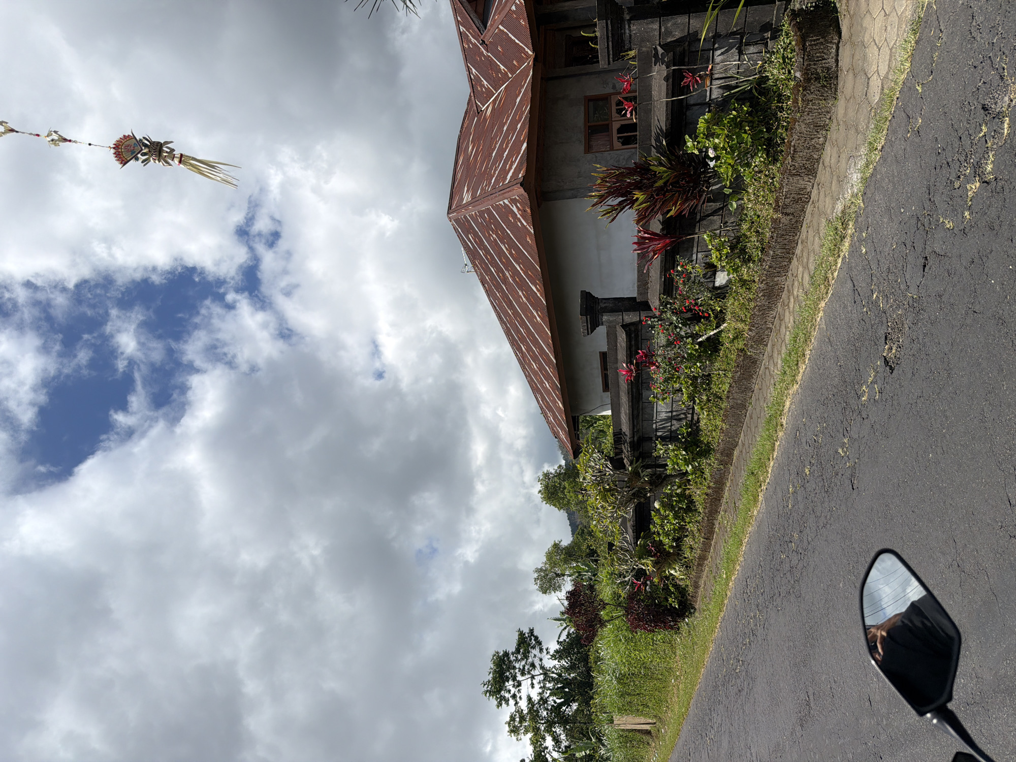 In Kintamani, Indonesia, a view from the perspective of a rider on a motorcycle reveals a weathered asphalt road. To the right, a stone wall borders a property with a single-story building topped with a rusty, corrugated metal roof. Lush green vegetation and vibrant red plants are visible in front of the wall. Above the scene, a decorative Balinese offering hangs suspended in the cloudy sky. The motorcycle's black rearview mirror reflects the sky and the rider's hand, holding a phone.