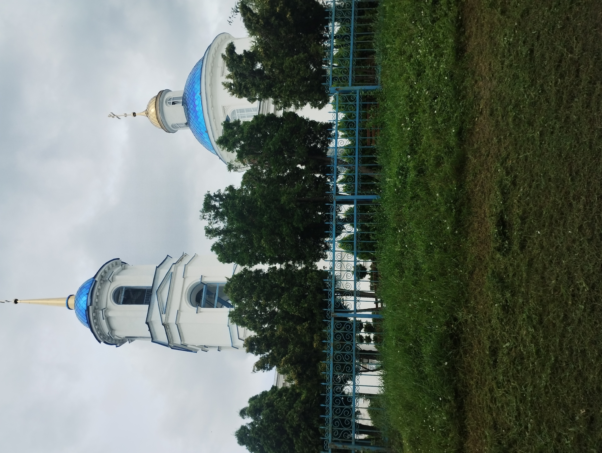 A low-angle, full-shot photograph captures an exterior view of the Transfiguration Cathedral in Sumy, Ukraine, on a cloudy day. The white, neoclassical church features a prominent bell tower topped with a blue dome and a golden spire, and a larger blue dome with a golden cap and cross. A bright blue, ornate metal fence encloses the property. Lush green grass and trees surround the church, with the foreground showing freshly cut grass. The sky is overcast with gray clouds.