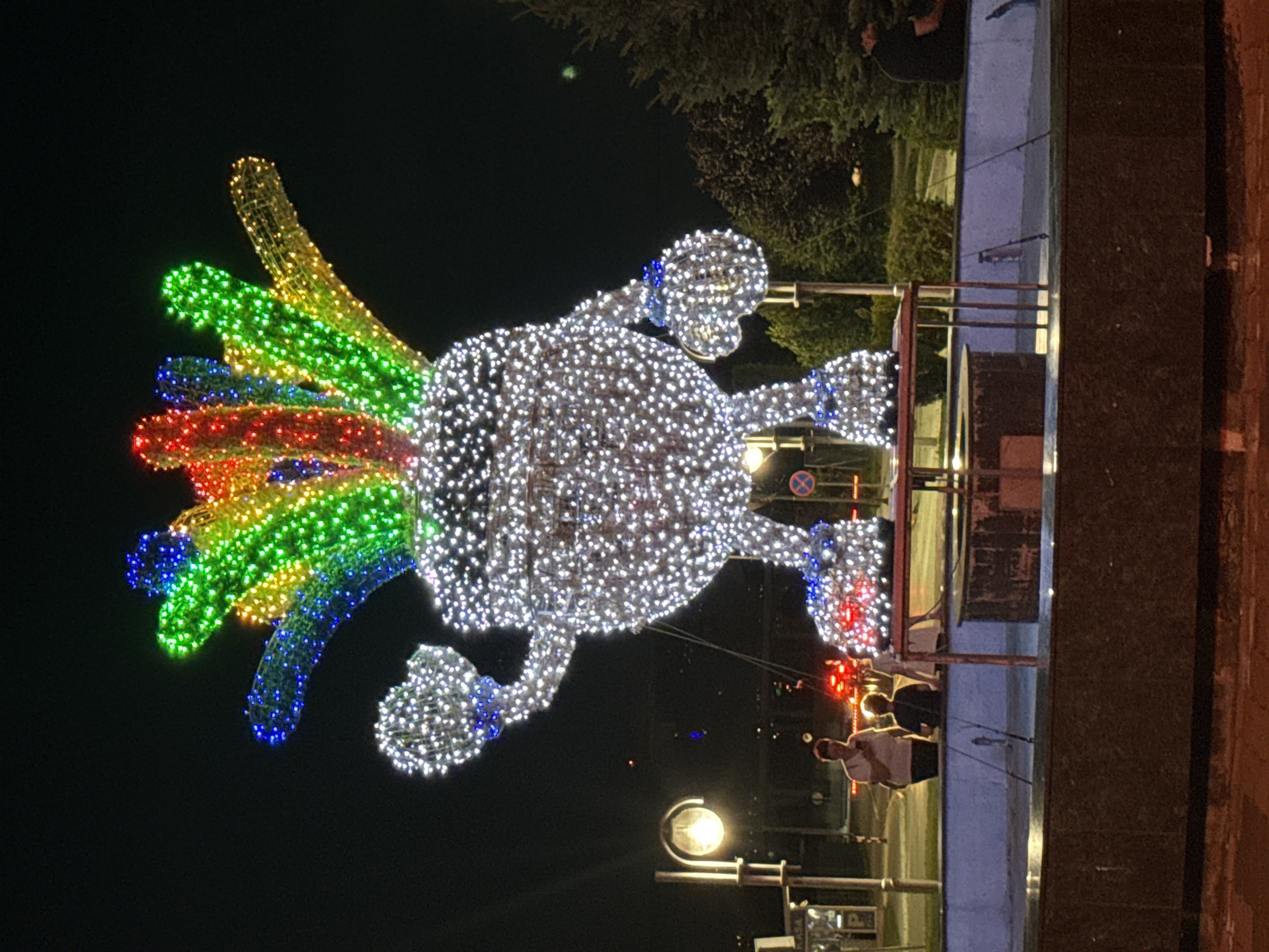 The image shows a large, illuminated sculpture in the city of Ilidža, Bosnia and Herzegovina, at night. The sculpture is of a cartoonish character made of white lights, with a headdress of colorful lights in red, blue, green, and yellow. The character has round arms and legs, and it appears to be standing on a base surrounded by a fountain. Two people, an adult and a child, are standing in the background near a streetlamp, and another person is sitting in the distance. The environment appears to be an urban outdoor setting with trees and streetlights visible.