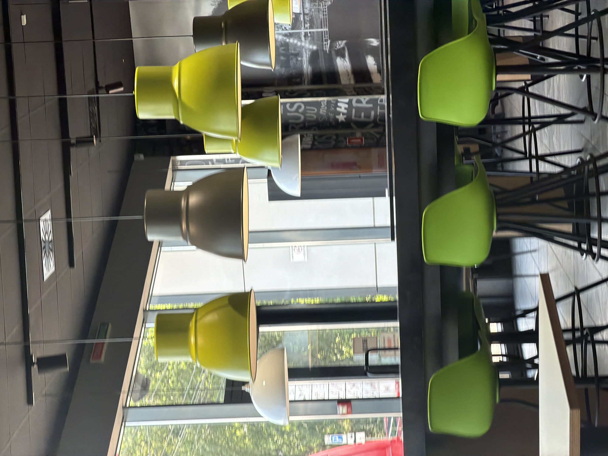 The image depicts an interior scene, likely a restaurant or cafe, with a modern aesthetic. The foreground features a section of a long, dark-colored counter or bar. Several bright green bar stools with black metal legs are arranged around it, with one prominent stool on the left and another on the right of the frame. A light-colored table with a wooden edge is partially visible in the lower left corner.
Suspended from the ceiling are multiple pendant lights. The dominant light fixtures are large, bell-shaped lamps in a vivid lime green. Some of these green lamps are in focus, while others are slightly blurred. Interspersed among them are two darker, gray bell-shaped lamps and one white bell-shaped lamp. The ceiling itself appears to be a dark grid system, with recessed lighting elements visible.
Behind the counter, a large window offers a view of the outdoors. Lush green trees and foliage are visible through the glass, suggesting a daytime setting. The window frames are dark and possibly metallic. Reflections are visible on the window panes and the black surfaces in the background, showing more of the interior lighting and some indistinct imagery on a wall, possibly a mural or poster with text.
The overall atmosphere is contemporary and casual, characterized by the bright, bold color of the stools and some of the light fixtures, contrasted with the dark furniture and ceiling. There are no people visible in the image. The scene does not contain any specific markers that would identify its location in Kyiv, Ukraine.