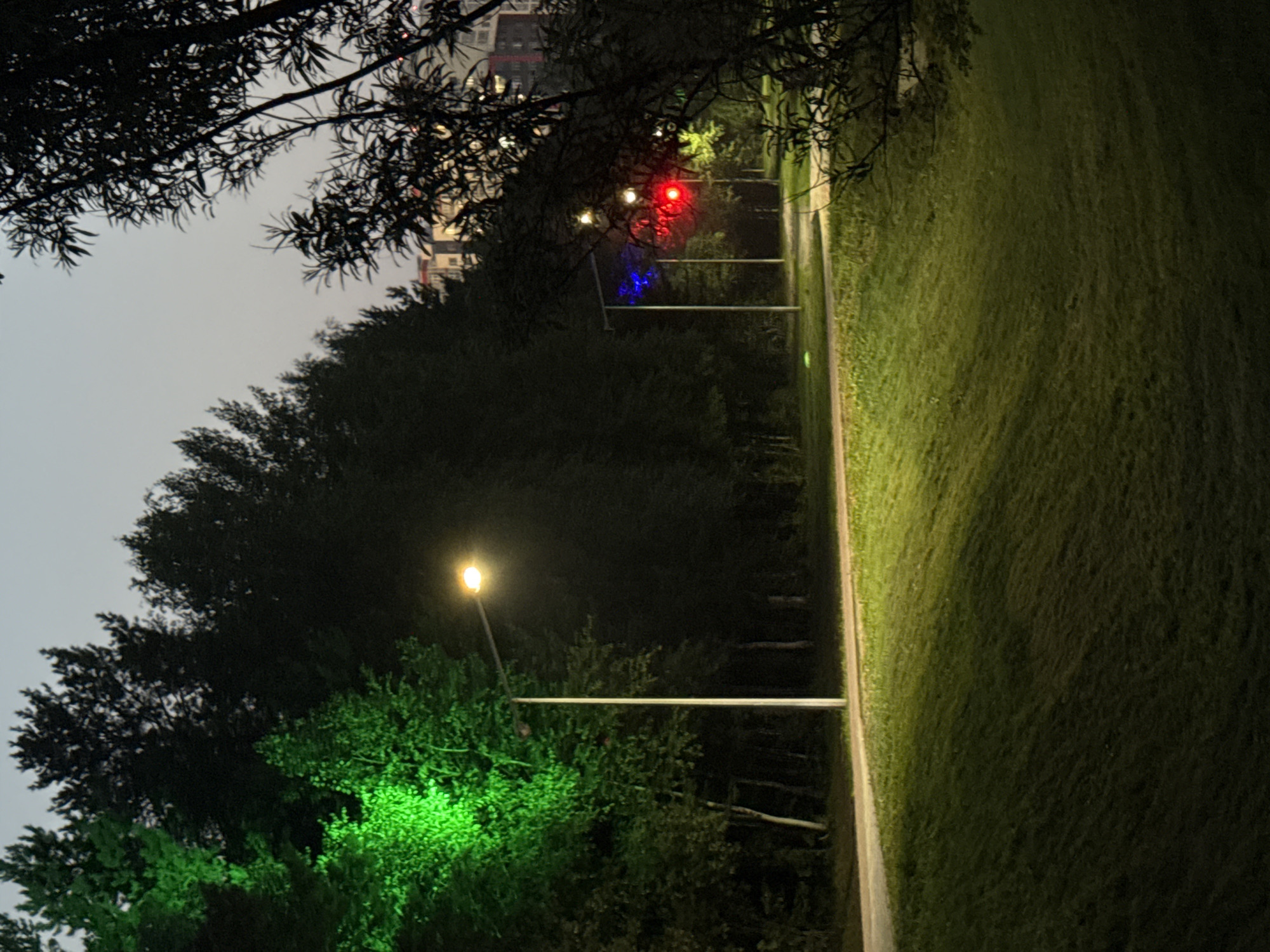 In an outdoor park setting, a pathway winds through a grassy area bordered by trees. The sky is a dusky gray, suggesting it is either dawn or dusk. Multiple streetlights illuminate the scene. One streetlight on the left casts a vibrant green light onto the surrounding trees. Further down the path, two more streetlights are visible; one emits a bright red light and the other a blue light. In the background, a few residential buildings are visible, hinting at the park's proximity to an urban area. The overall atmosphere is calm and tranquil.