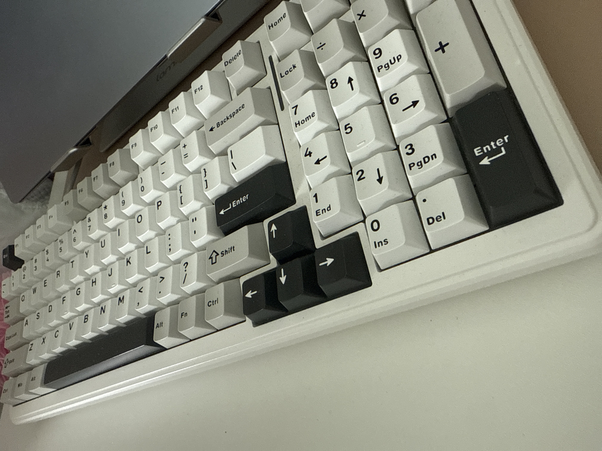 The image shows a white computer keyboard with black keycaps on some of the keys. The keyboard is resting on a white surface, possibly a desk. Some of the keycaps are labeled with "Enter", "Shift", and arrow symbols. The keyboard appears to be a full-size keyboard with a number pad. Part of a laptop is also visible in the upper left corner of the image.