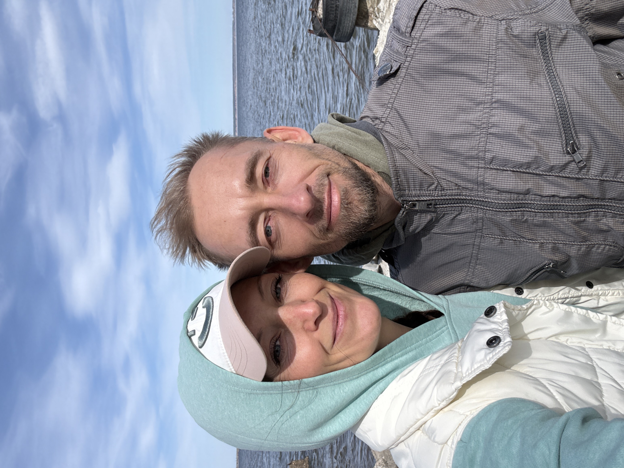 A man and a woman are captured in a bright, outdoor selfie-style portrait. The image is oriented vertically, showing the subjects smiling happily.
The man, positioned slightly behind and to the left of the woman, has short light brown hair and a neatly trimmed beard. He is wearing a grey jacket with a subtle plaid pattern and a visible zipper on the chest pocket, layered over what appears to be a greenish hoodie. He gazes directly at the camera with a warm smile.
The woman is in the foreground, also smiling towards the camera. She wears a light pink baseball cap with a white front panel featuring a prominent dark "C" logo. Her attire includes a light turquoise hoodie, and she has a white puffer vest over it, with black snap buttons visible.
Behind the couple, a wide expanse of water stretches out, indicating a large river or lake, consistent with the location context of Ulyanovsk, Russia, on the Volga River. The water surface shows small ripples. Above the water, a clear blue sky is visible with scattered, wispy white clouds. On the far right, behind the man, some rocky or concrete shoreline is discernible, with what appears to be a dark, old tire or similar object partially submerged or resting on the bank. The overall atmosphere is cheerful and relaxed, suggesting a pleasant day spent outdoors by the water.