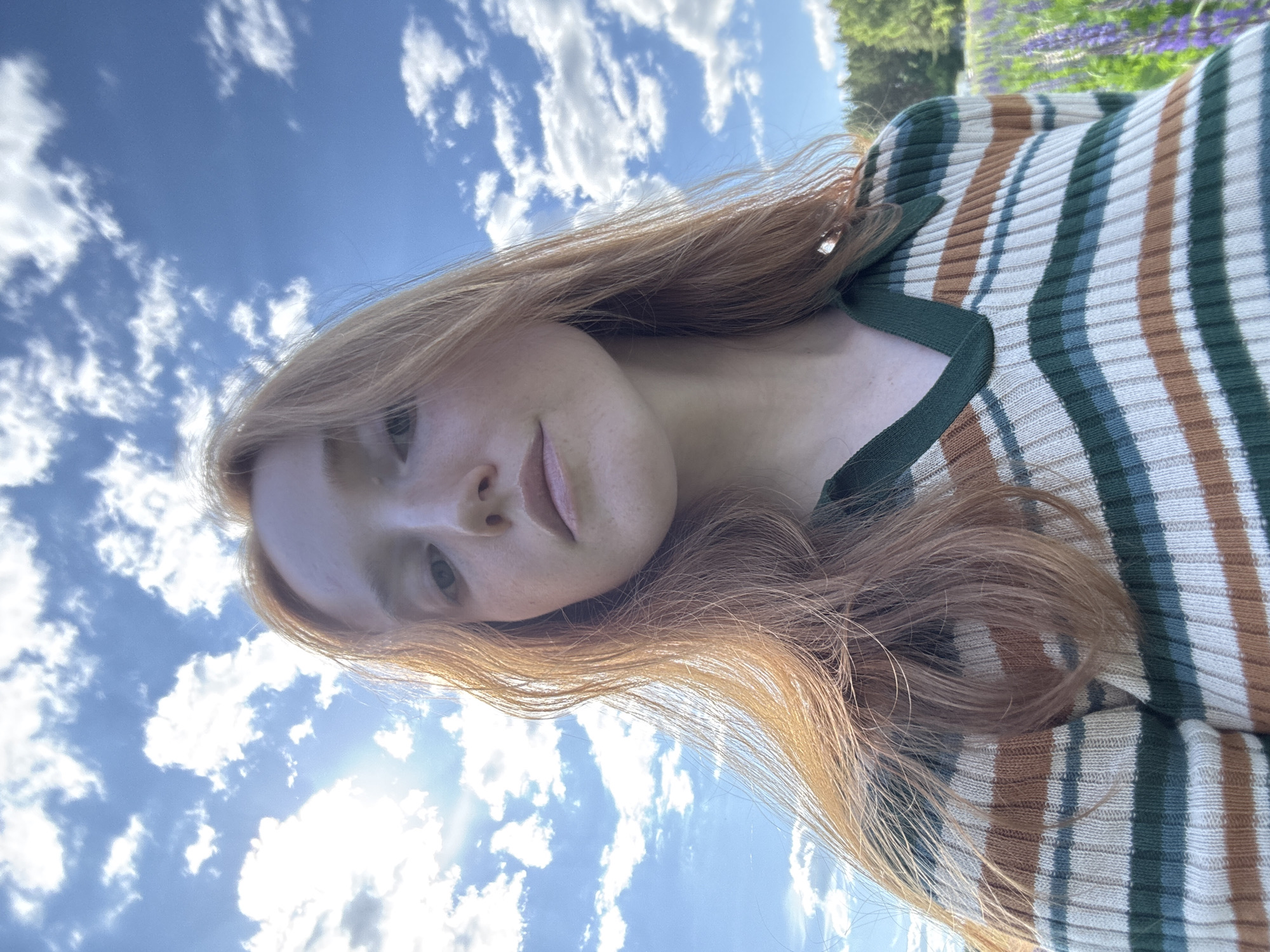 A young woman is depicted in a close-up, outdoor self-portrait, identified as being in the city of Chaikovsky, Russia. Her face is centered, showing light skin with visible freckles and a neutral expression as she looks directly at the camera. Her long, reddish-brown hair flows over her shoulders and chest, illuminated by strong sunlight originating from the upper left, which also creates a significant lens flare in that area of the frame. She is wearing a ribbed knit V-neck sweater with horizontal stripes in dark green, light teal, cream, and brown hues. The background consists primarily of a bright blue sky filled with numerous scattered white cumulus clouds. In the lower right, a field of tall purple flowers, consistent with lupines, is visible, bordering a line of dense green trees in the distant background.