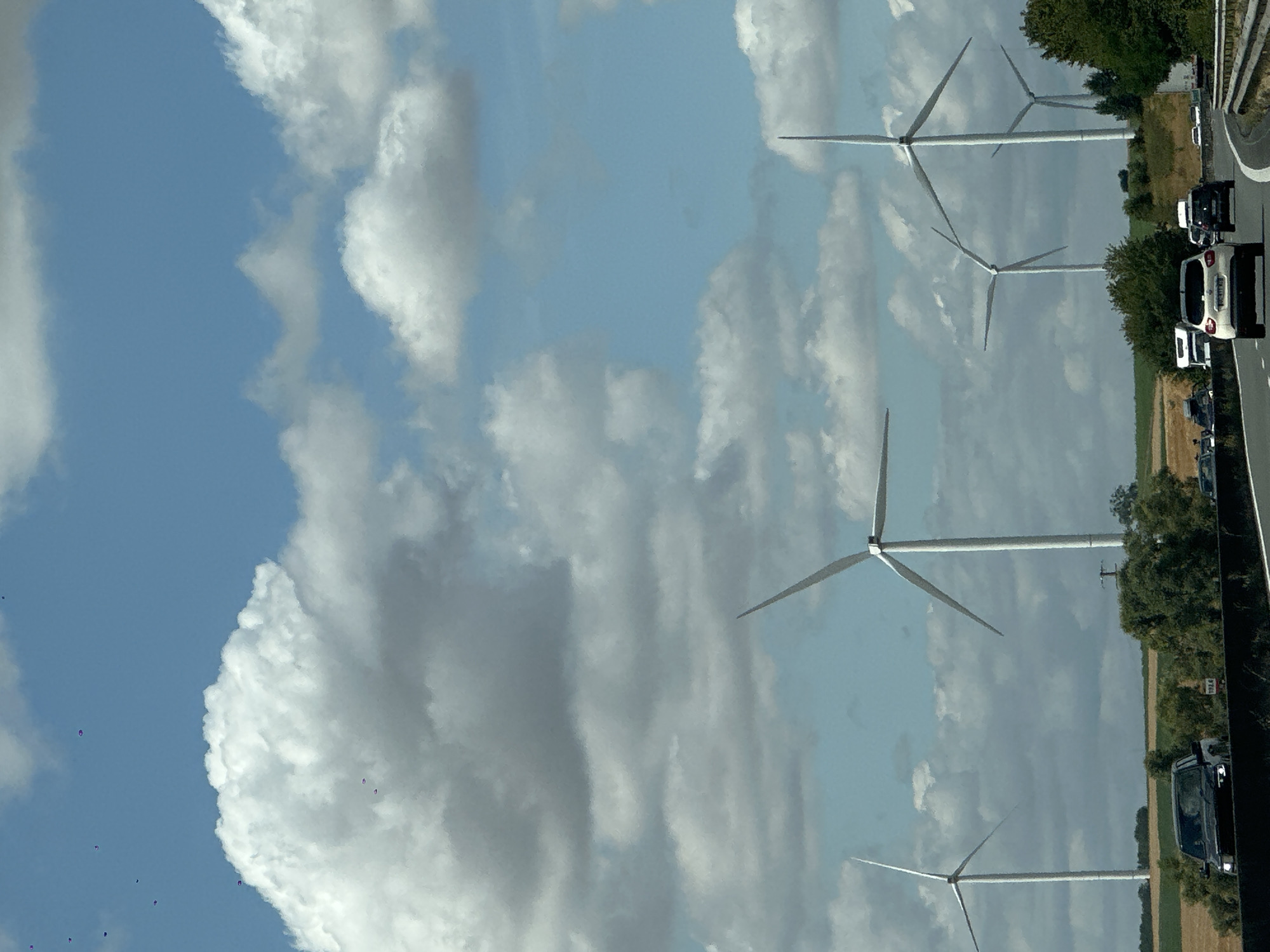 This is an outdoor, eye-level, vertical, daytime photograph taken from a vehicle on a highway. The scene depicts a landscape with multiple wind turbines in various stages of rotation against a cloudy blue sky. The foreground shows a highway with several cars and trucks, including a white SUV in the center lane, a black SUV on the left, and a truck on the right. The middle ground features rolling hills with fields of dry grass and scattered trees. The background is dominated by the imposing structures of the wind turbines, with the sky above filled with puffy white and grey clouds. The location is identified as Nouvion-le-Comte, France.