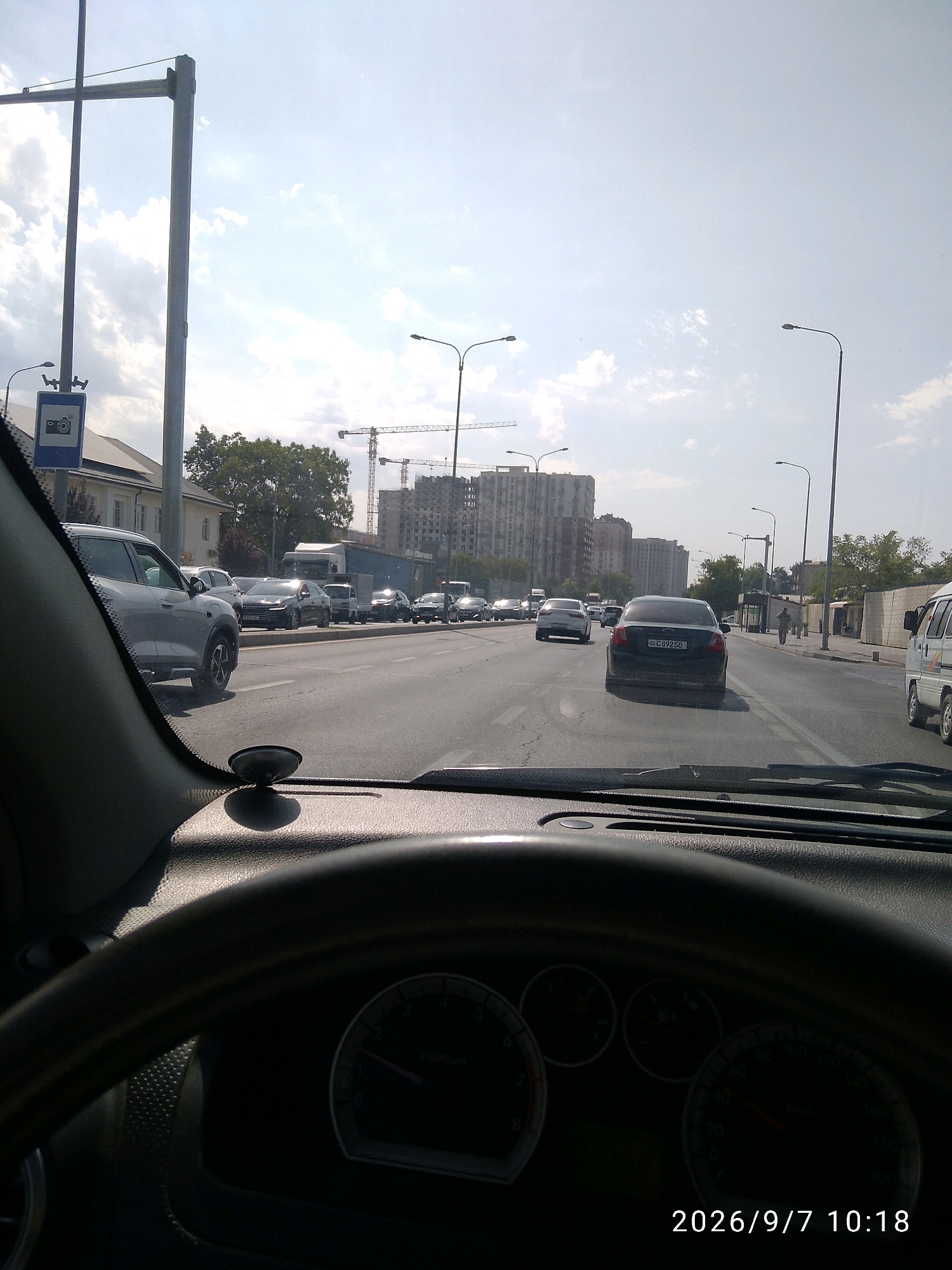 A driver's perspective shows a traffic jam on a sunny day in Tashkent, Uzbekistan. The view is from inside a car, looking out through the windshield. The steering wheel and dashboard are visible in the foreground. Cars are stopped in multiple lanes on a wide asphalt road. On the left, a white SUV is in the foreground. Further down the road, a black sedan with license plate "DC892SB" is prominent. In the background, tall residential buildings under construction and cranes suggest ongoing urban development. The sky is bright with scattered clouds. A traffic sign with a camera icon is visible on a pole to the left. The date and time stamp "2026/9/7 10:18" is superimposed on the image.