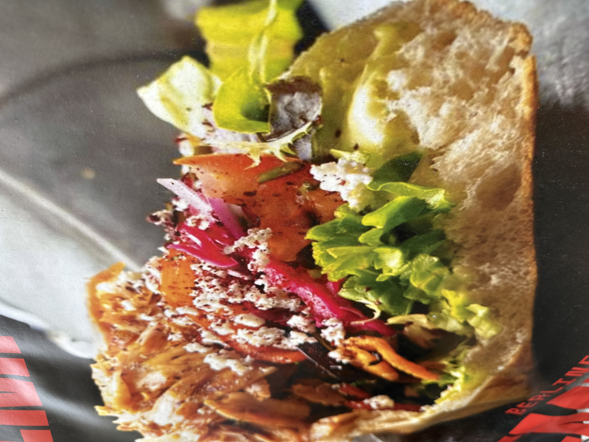 The image depicts a Döner kebab in a bread pocket. Ingredients include sliced meat, tomatoes, red onions, lettuce, shredded carrots, and a white sauce or crumbled cheese. The kebab appears to be presented in a paper wrap. The location in the city of Paris, France, cannot be verified from the image.