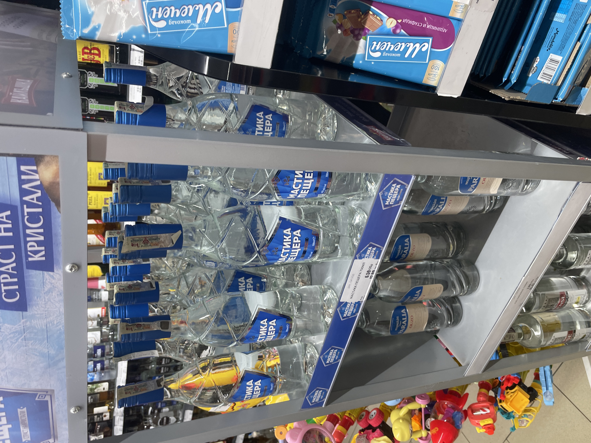 This image, captured indoors in what appears to be a supermarket or retail store in Ihtiman, Bulgaria, showcases shelves stocked with various products. The photo is rotated 90 degrees clockwise, making the shelves appear oriented vertically.
The central focus is on multiple rows of clear glass bottles, predominantly featuring "МАСТИКА ПЕЩЕРА" (Mastika Peshtera), a Bulgarian anise-flavored alcoholic beverage, identifiable by its distinctive blue labels and caps. These bottles are marked "700ml e". Adjacent to these, other bottles labeled "PARALIA OUZO" (Ouzo Paralia) are also visible. A price tag clearly shows "МАСТИКА ПЕЩЕРА 700МЛ" priced at "6.59 лв/бр" (Bulgarian Leva per piece).
Above these spirits, stacks of blue-packaged chocolate bars are displayed. Text on these packages includes "Млечен шоколад" (Milk chocolate), "ЛЕШНИЦИ И СТАФИДИ" (Hazelnuts and raisins), and the brand "Своге" (Svoge), along with "100г". Partially visible on a higher shelf are bottles of "JB" whiskey.
A prominent shelf-edge sign on the left features Cyrillic text, reading "СТРАСТ НА КРИСТАЛИ" (Passion of Crystals) and "КАЙЛЫШКА" (Kailashka). On the lowest shelf visible, a collection of colorful children's toys, in yellow, red, and pink hues, suggests a mixed retail environment. No people are present, and the scene depicts a static product display.