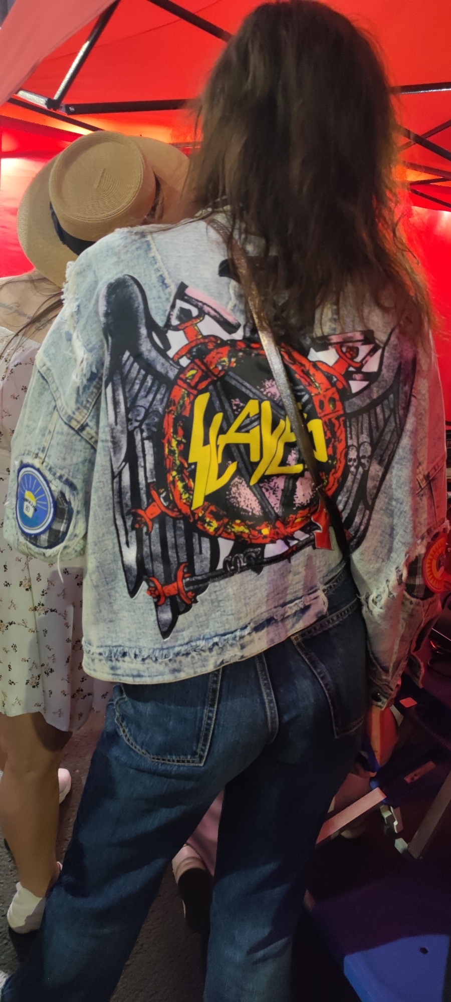 The image depicts the backs of two individuals standing under a red canopy, suggesting an outdoor or semi-outdoor event in low light. The central figure, viewed from the back, has long, dark brown, wavy hair and wears a distressed, light blue denim jacket with a large, elaborate "Slayer" band logo prominently displayed. The logo features a winged sword, an inverted pentagram, and the band's name in yellow script, all within a circular design with red and black elements. The individual also wears dark blue jeans. A dark shoulder strap of a bag crosses their back. On the left sleeve of the jacket, a circular patch with blue, white, and yellow elements, including the number "80", is visible. A red and white plaid patch is partially visible on the right sleeve. To the left, another person is partially visible, wearing a straw hat with a dark band and a light-colored dress with a subtle floral pattern. Their feet, clad in white sneakers, are visible on the dark ground. Additional legs and feet of other people are visible at the bottom of the frame, indicating a gathering. The overhead structure is a red fabric canopy supported by black metal beams. The ambient lighting is predominantly red from the canopy, creating a warm, dim atmosphere.
