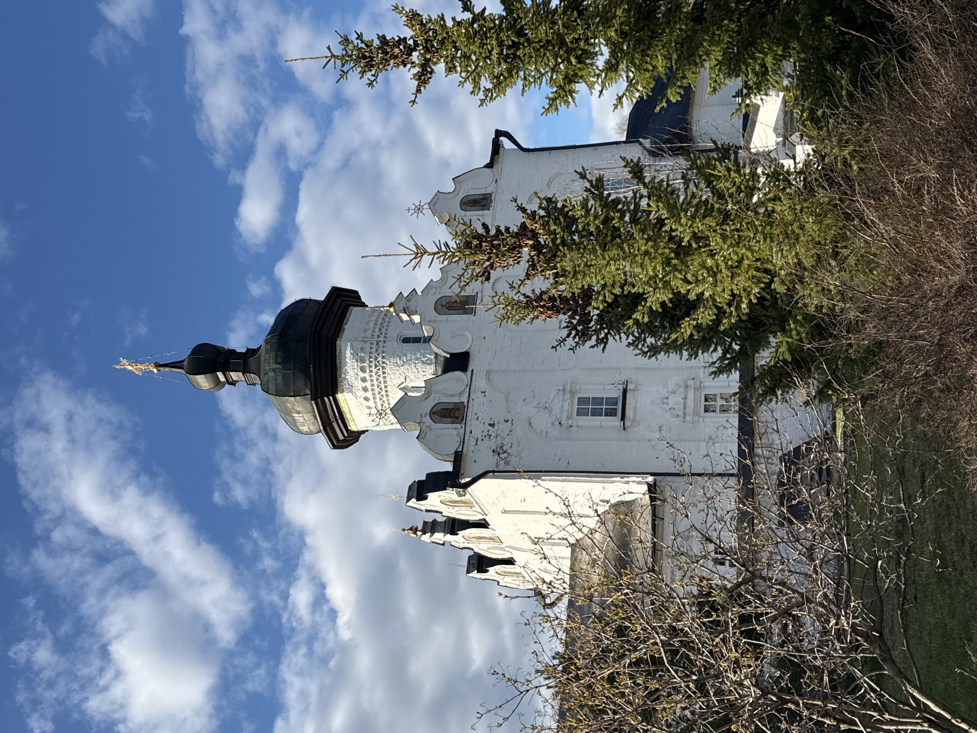 The image depicts a white building with a black dome and a golden spire, partially obscured by trees. The building is likely part of a monastery complex due to its architectural style. The sky above is blue with scattered white clouds. The trees in the foreground are bare, suggesting it is either late autumn or early spring. The building and the surrounding trees are located in the city of Sviyazhsk, Russia.