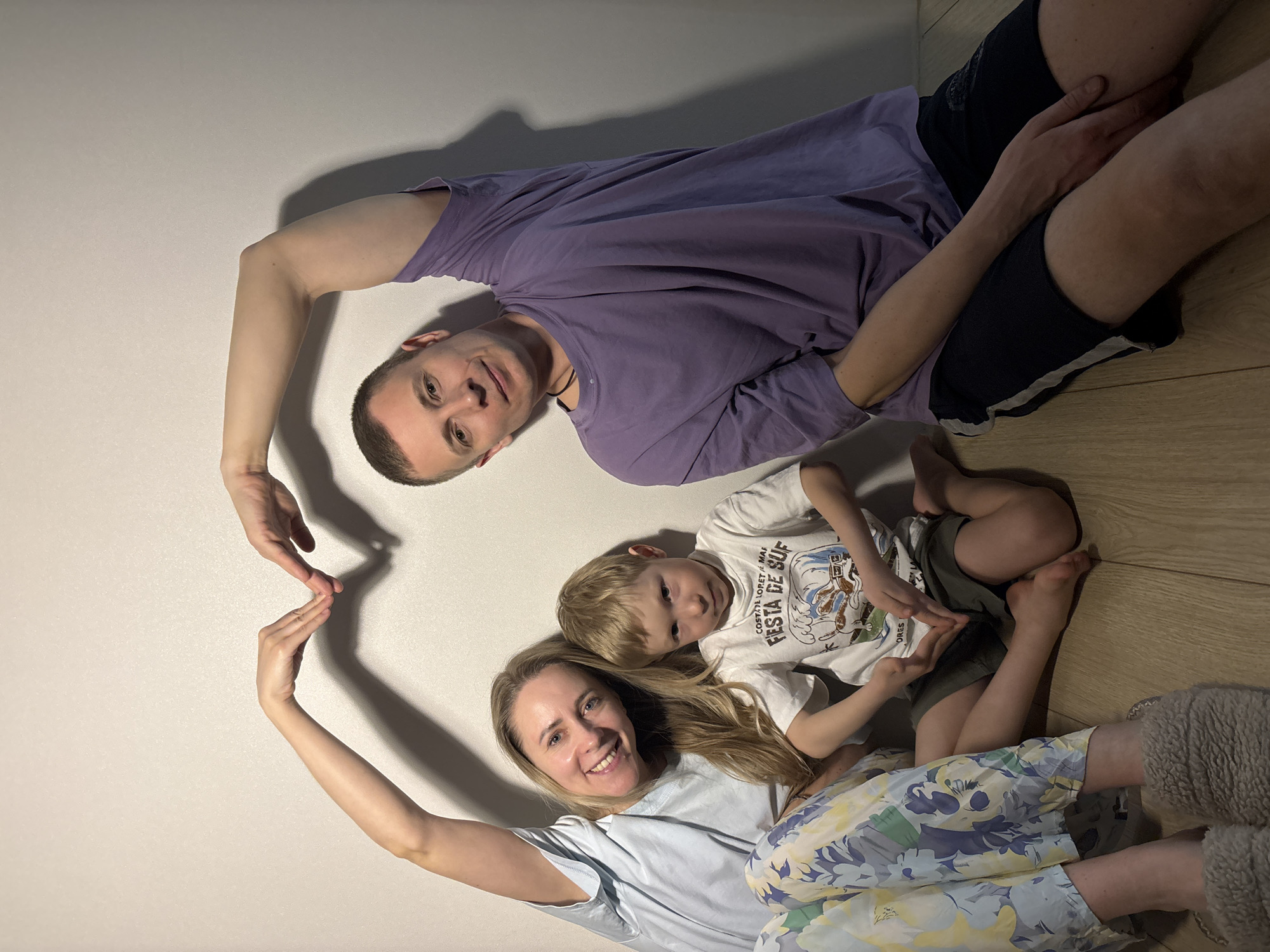 A family portrait, captured from an overhead perspective, shows a father, mother, and their young son lying on a light wooden floor against a plain white wall in a home in Chaikovsky, Russia. The man, on the right, wears a purple t-shirt and dark shorts, and looks directly at the camera. His left arm is bent to form the upper right curve of a large heart shape. The woman, on the left, wears a light blue t-shirt, floral pants, and grey fuzzy slippers, and smiles broadly at the camera. Her right arm is bent to form the lower left curve of the heart. Their fingertips meet to complete the large heart.
Their young son lies between them, leaning against the woman, wearing a white t-shirt and dark shorts, also smiling at the camera. He mirrors his parents by forming a smaller heart shape with his own hands. The setting appears to be indoors with artificial lighting, indicated by the distinct shadows cast on the floor and wall. The atmosphere is warm and joyful, highlighting family love and connection.
Visible text on the boy's white t-shirt reads: "COSTA LORET MAR FESTA DE SUF".